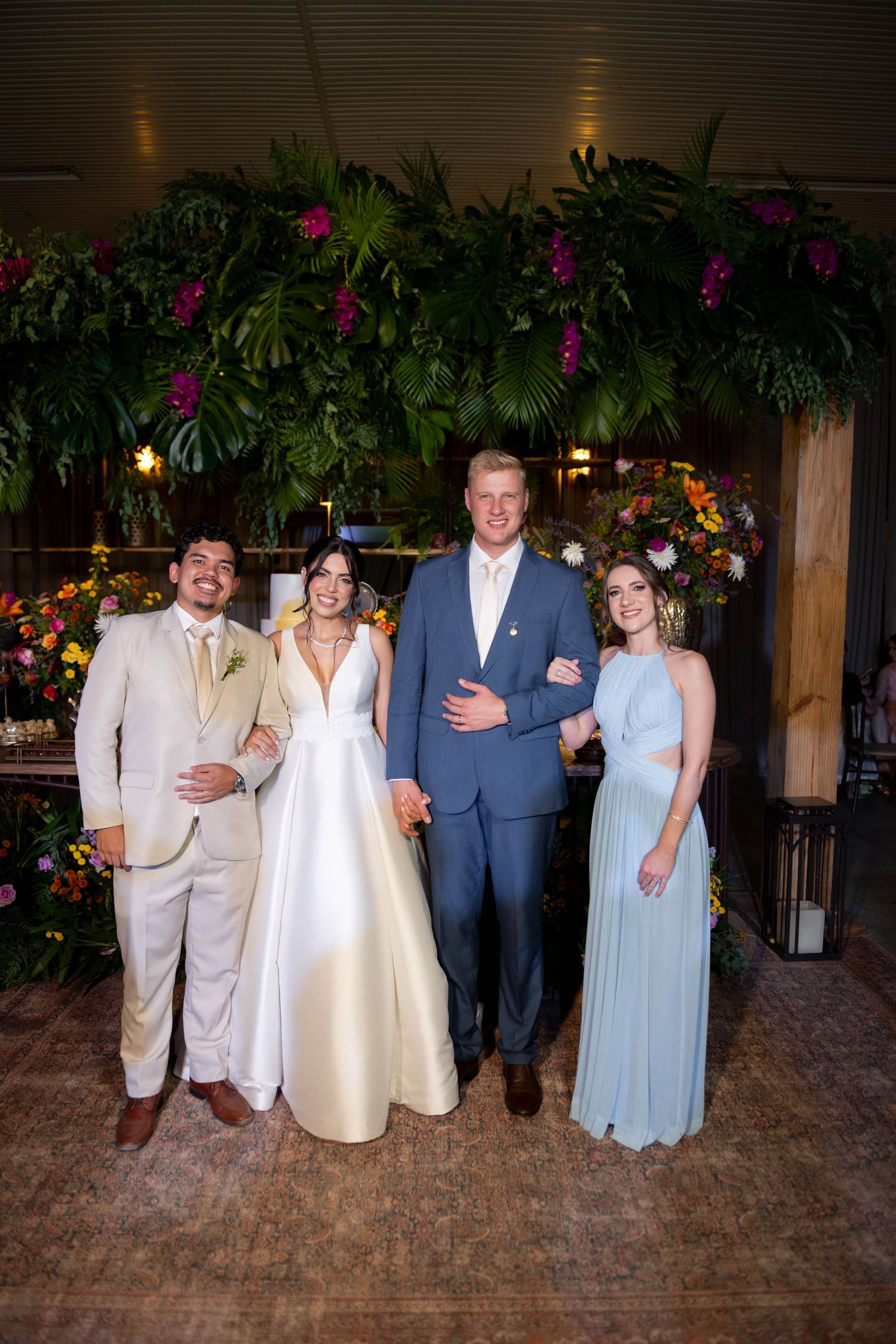 Wedding party of four posing indoors, bride in white gown, groom in blue suit, guests in cream and light blue formal attire.