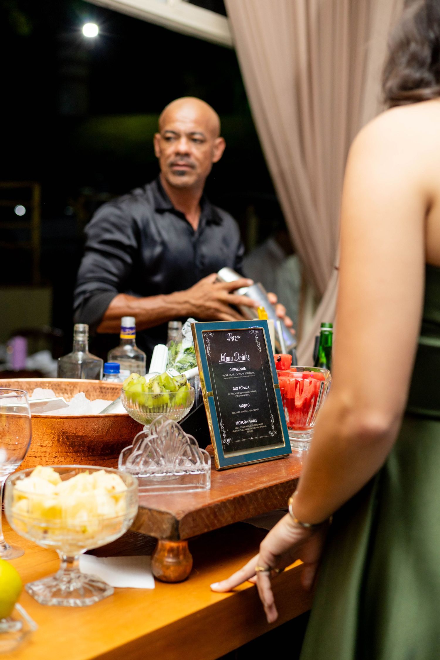 Bartender preparing cocktails at an elegant outdoor bar with fresh fruit garnishes and a drinks menu display.