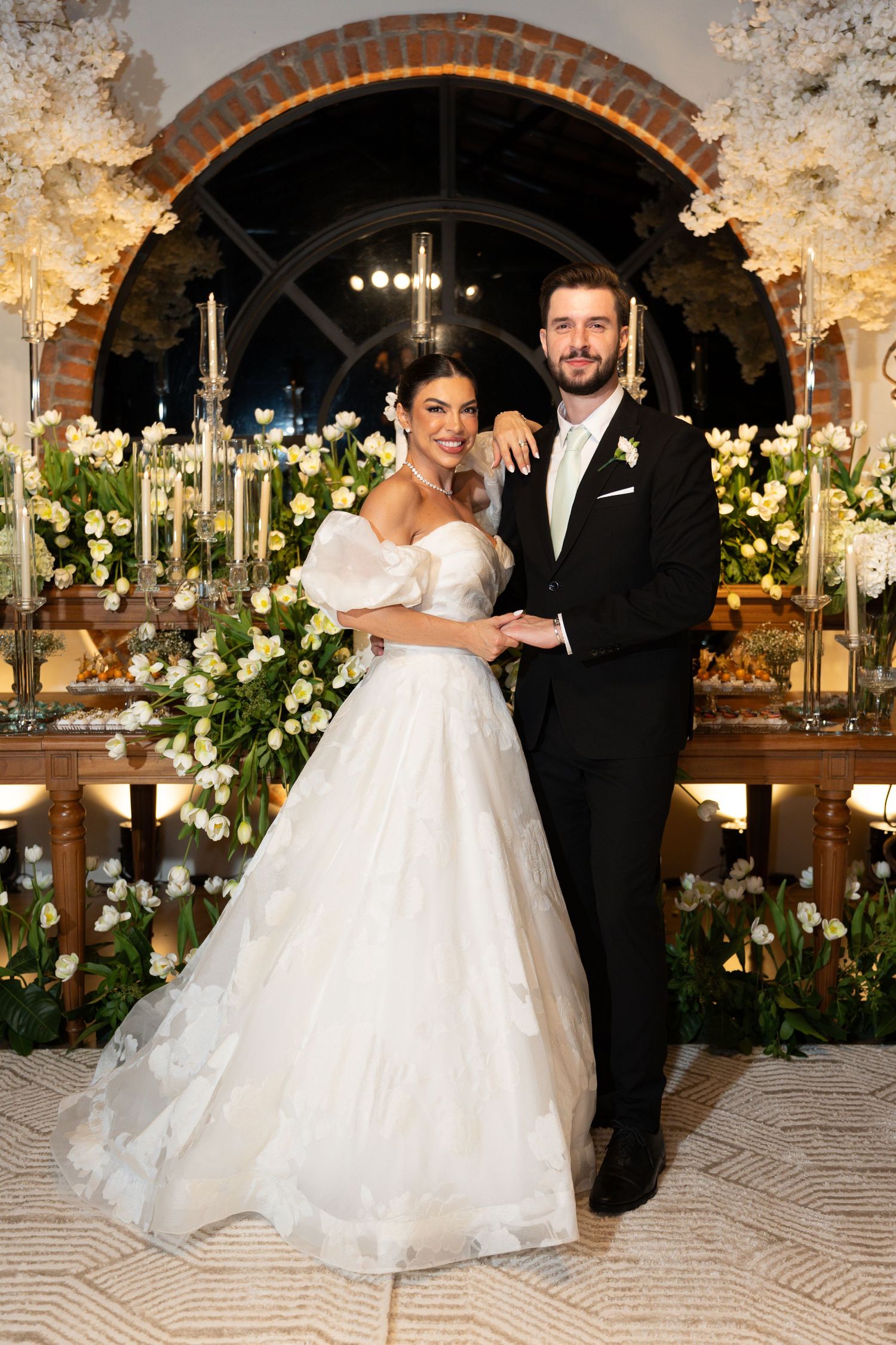 Bride in white off-shoulder ball gown and groom in black tuxedo pose together at elegant floral wedding reception.