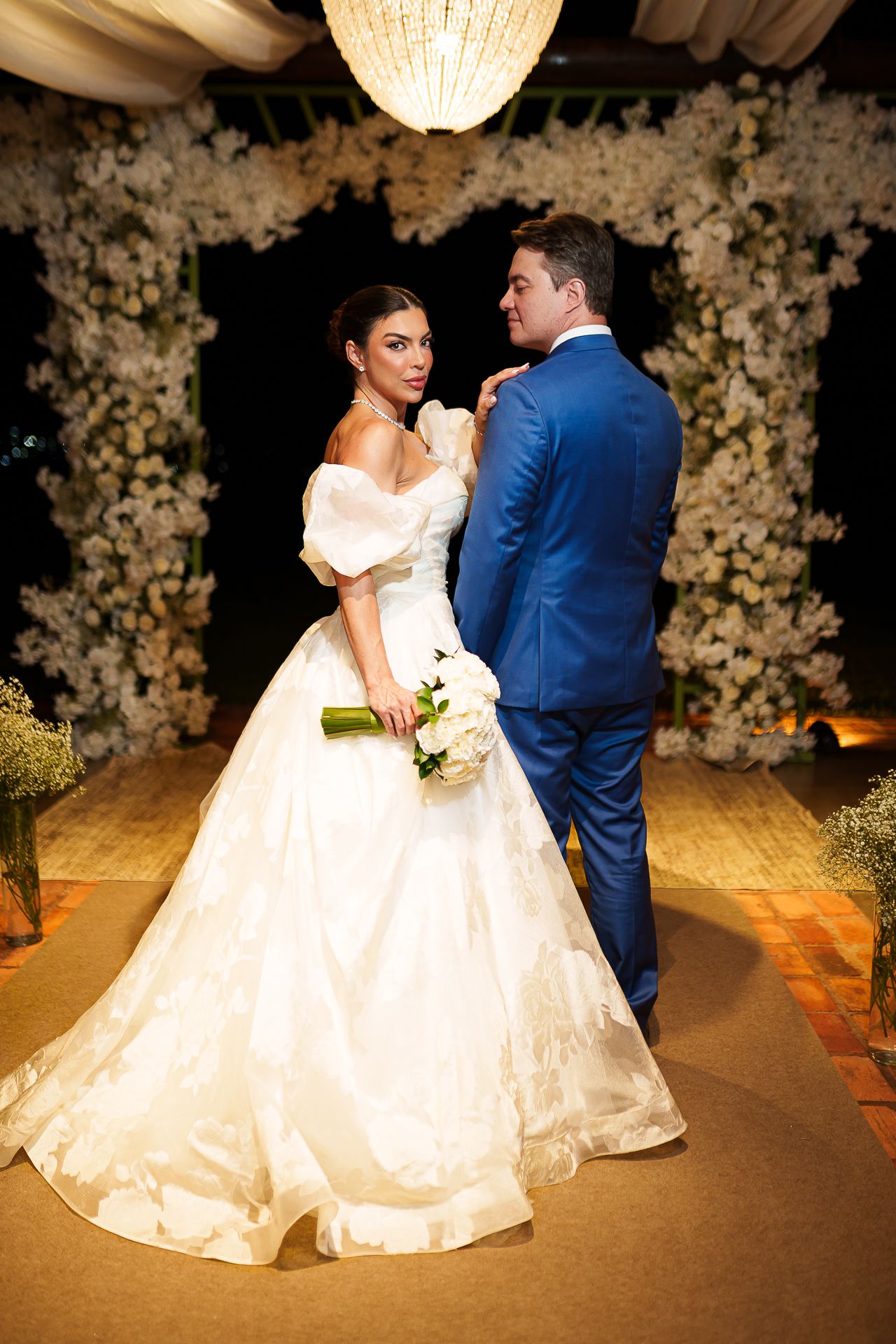 Bride in white ball gown holding bouquet stands with groom in blue suit before floral arch at elegant night wedding.