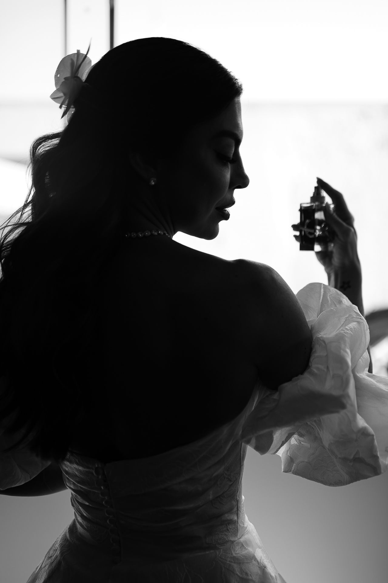 Black and white silhouette of a bride in a strapless gown holding perfume, with pearl necklace and flower in hair.