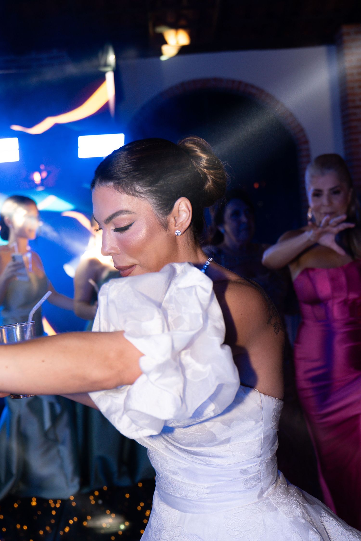 Bride in white ruffled gown dancing at wedding reception with guests in pink dresses visible in background.