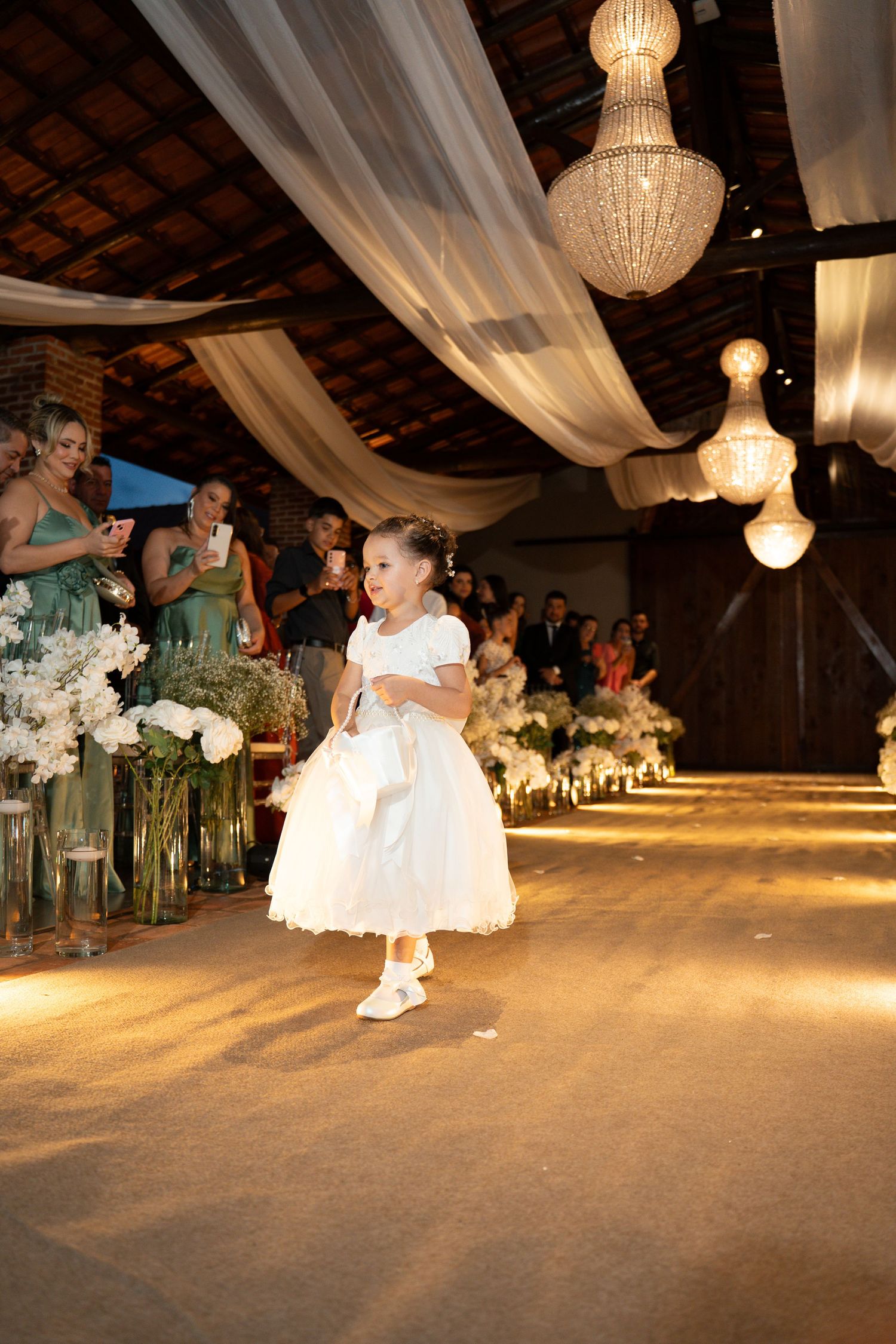 A flower girl in a white dress walks down a decorated wedding aisle with chandeliers and floral arrangements.