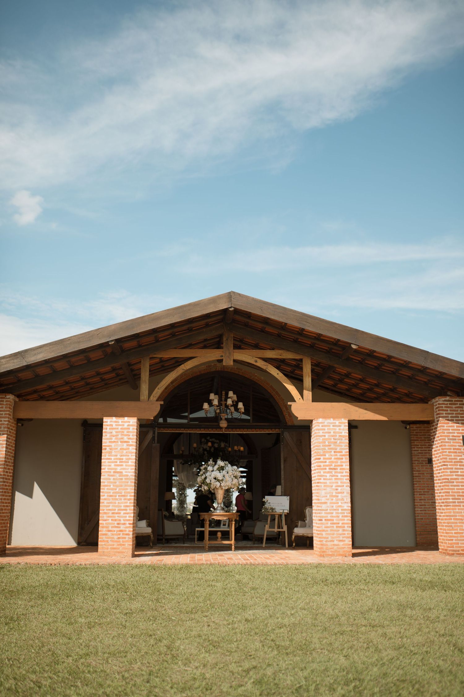 Rustic brick wedding venue with wooden arch entrance, floral decorations, and chandelier under blue sky.