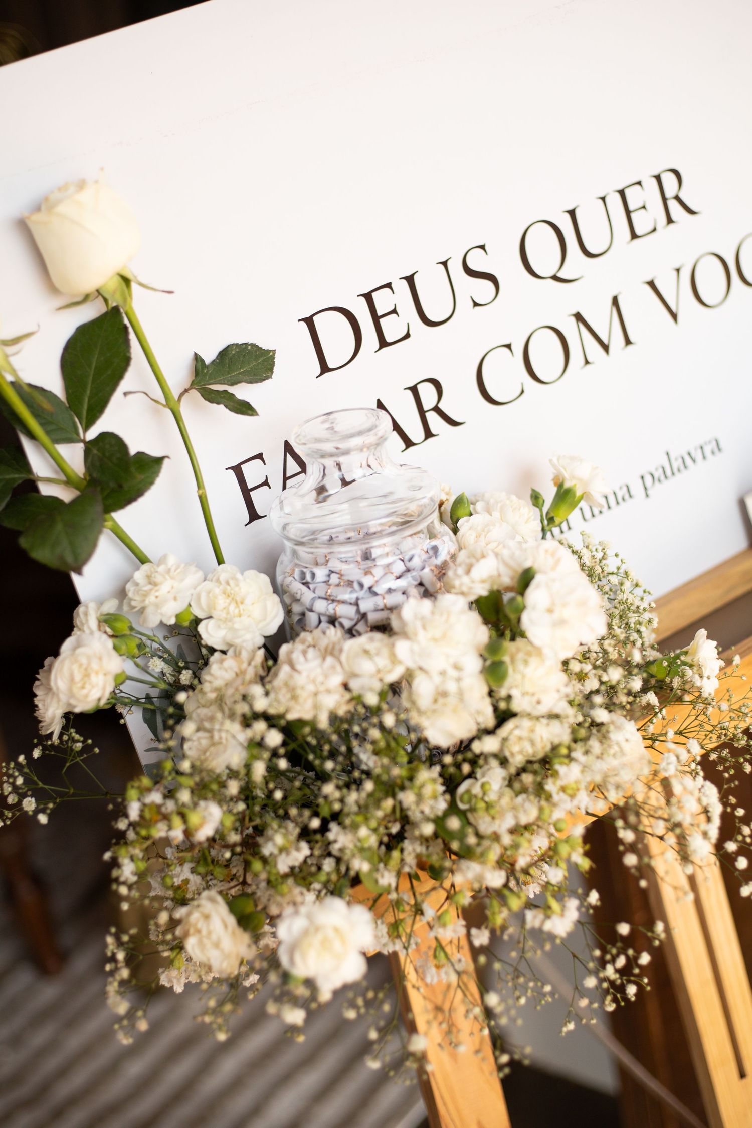 White floral arrangement with roses and baby's breath beside a Portuguese religious sign reading Deus Quer Falar Com Voce.