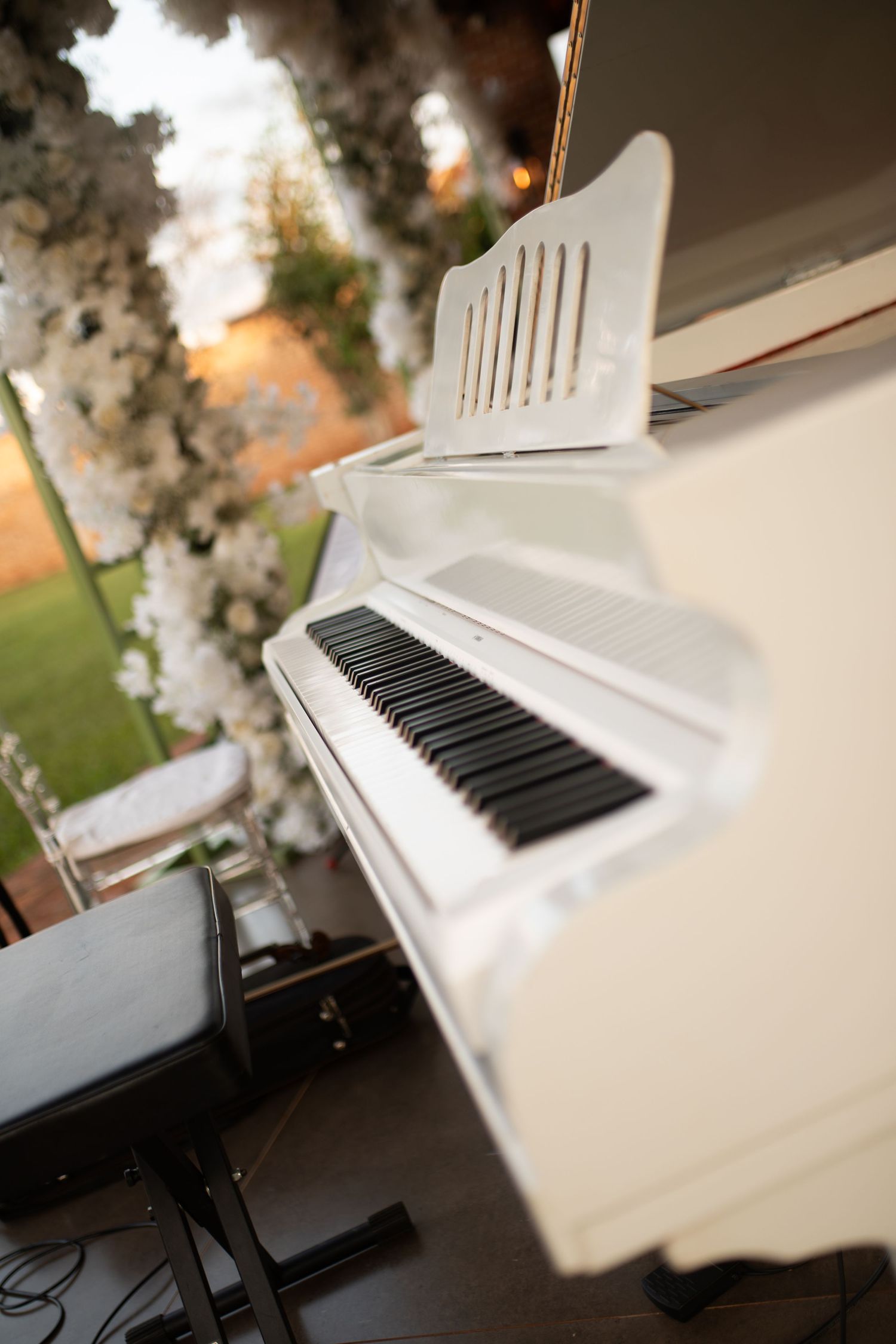 White grand piano with black keys outdoors at a wedding reception, surrounded by white floral arrangements.