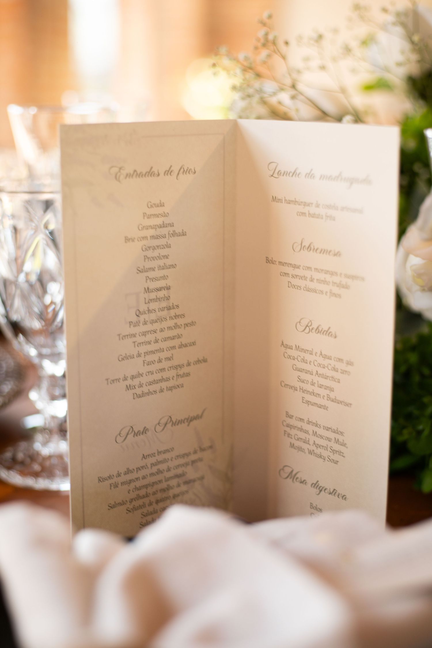 Elegant wedding menu card displayed on a table with crystal glassware and floral decorations in soft lighting.