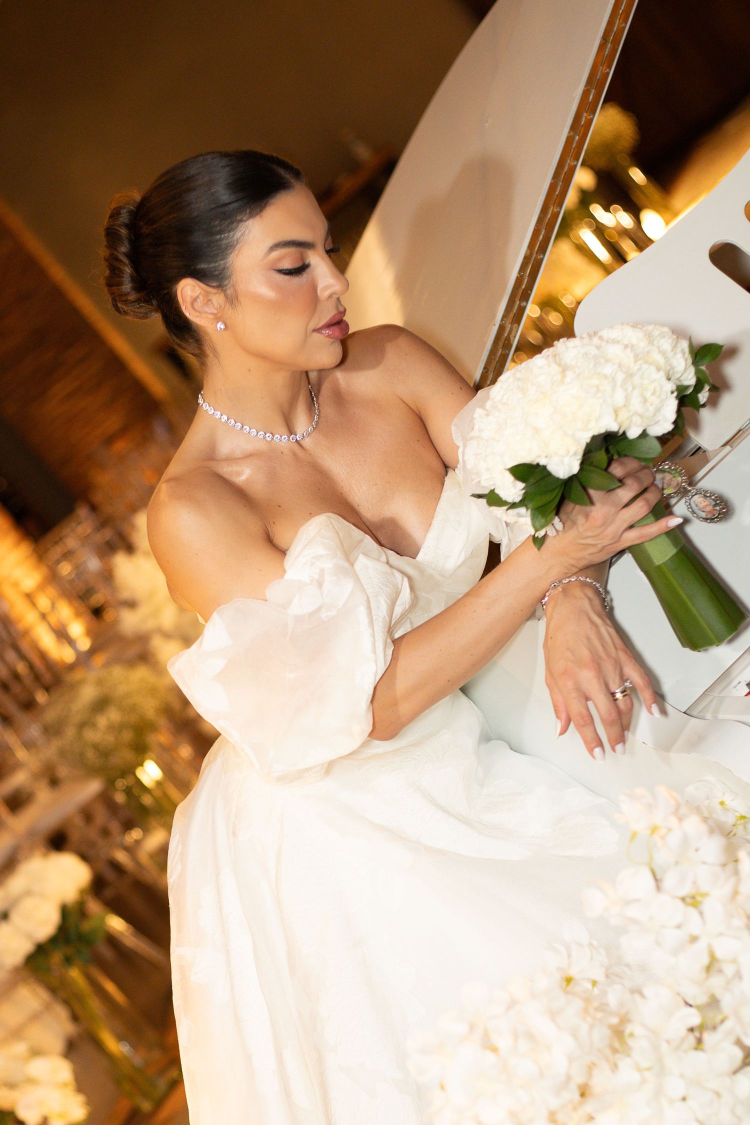 Elegant bride in white off-shoulder gown arranging white hydrangea bouquet, wearing diamond necklace at wedding reception.