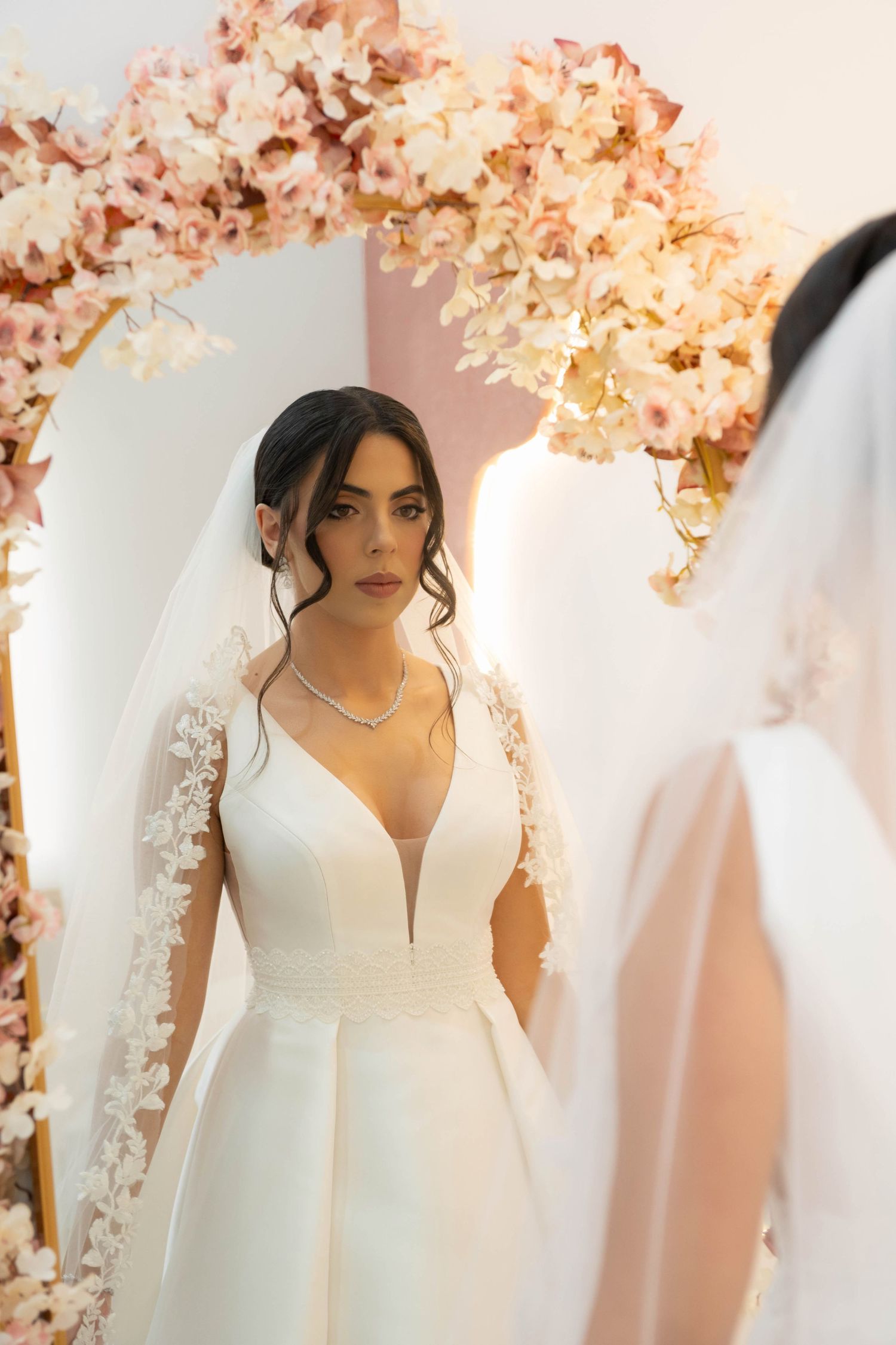 Bride in deep V-neck wedding gown with lace veil admires her reflection in mirror surrounded by pink floral arch.
