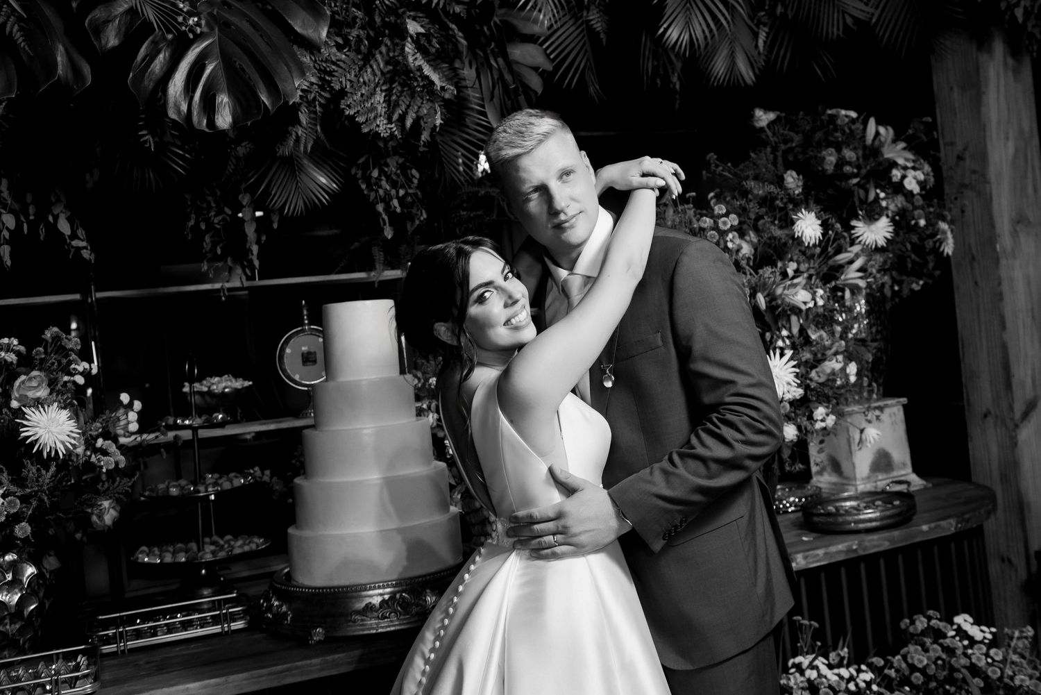 Black and white wedding photo of bride and groom embracing near tiered cake and lush floral arrangements.