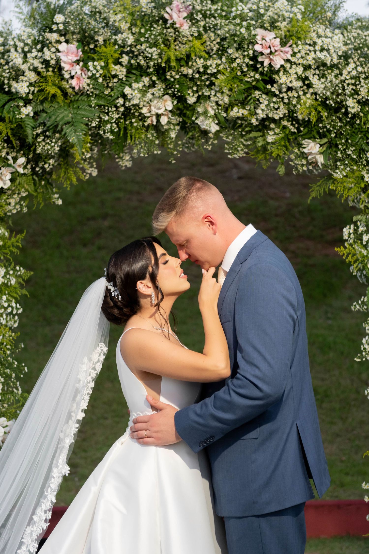 Bride and groom share a romantic moment under a floral arch adorned with white and pink blooms at an outdoor wedding.