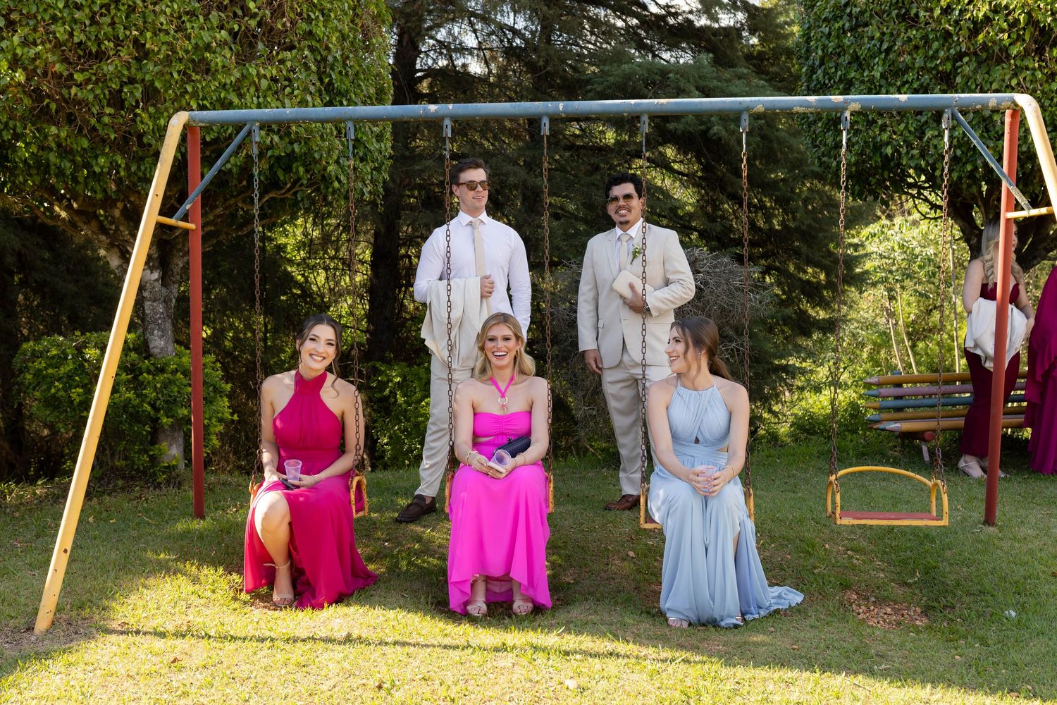 Five prom-dressed teens pose by a backyard swing set, three girls in colorful gowns and two boys in suits on a sunny day.