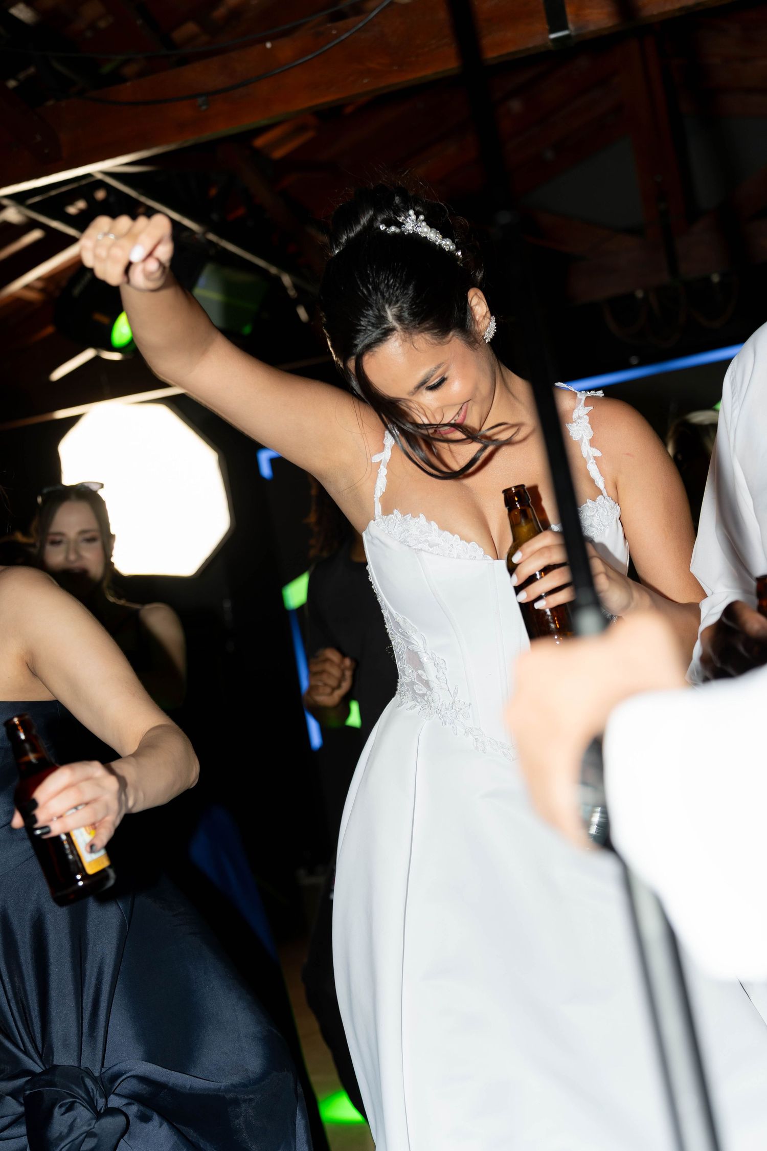 Bride in white wedding gown dancing energetically at reception, holding beer bottle with arm raised.