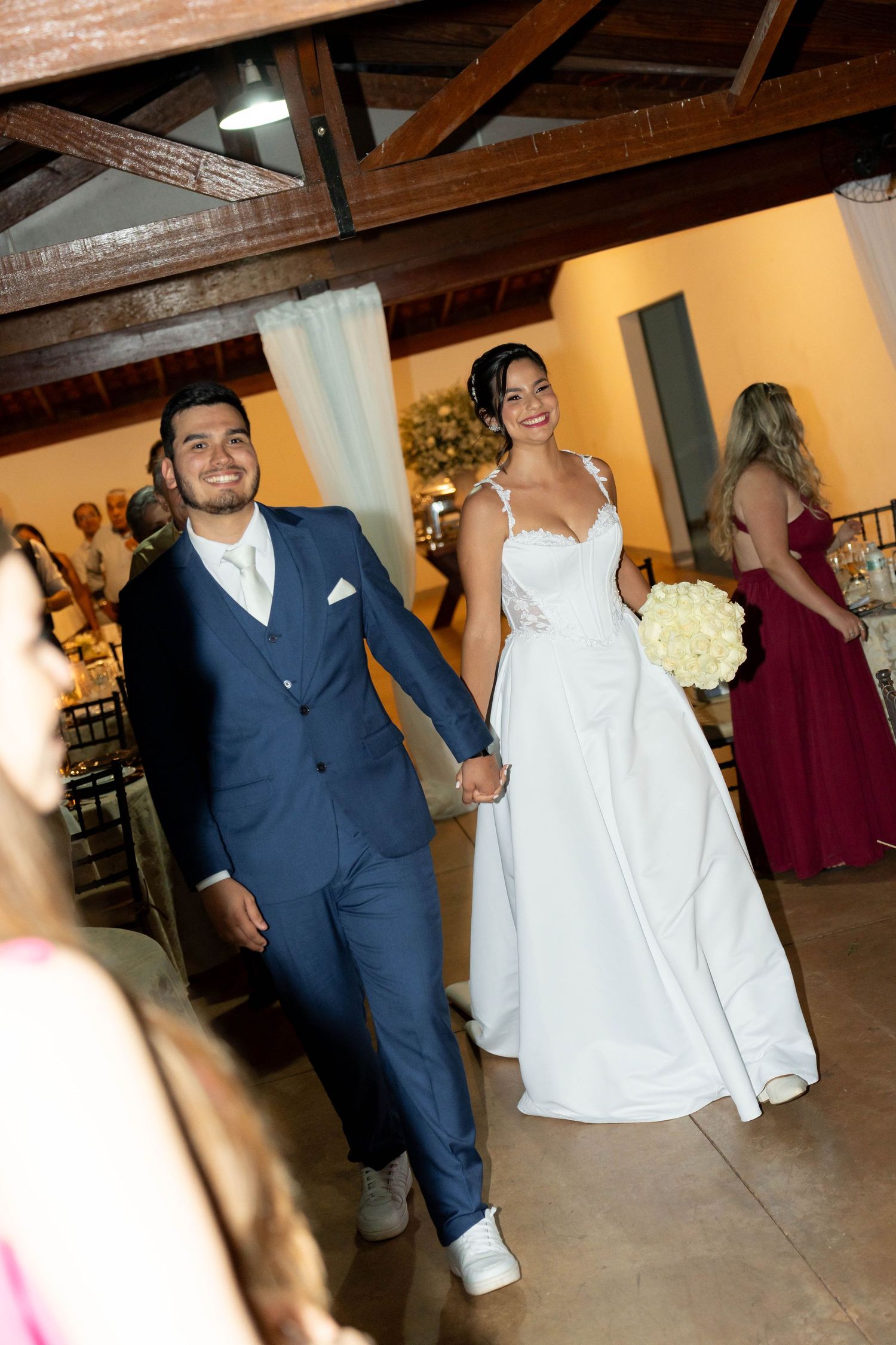Smiling bride and groom walk hand-in-hand at their wedding reception, bride holding white bouquet, groom in navy suit.