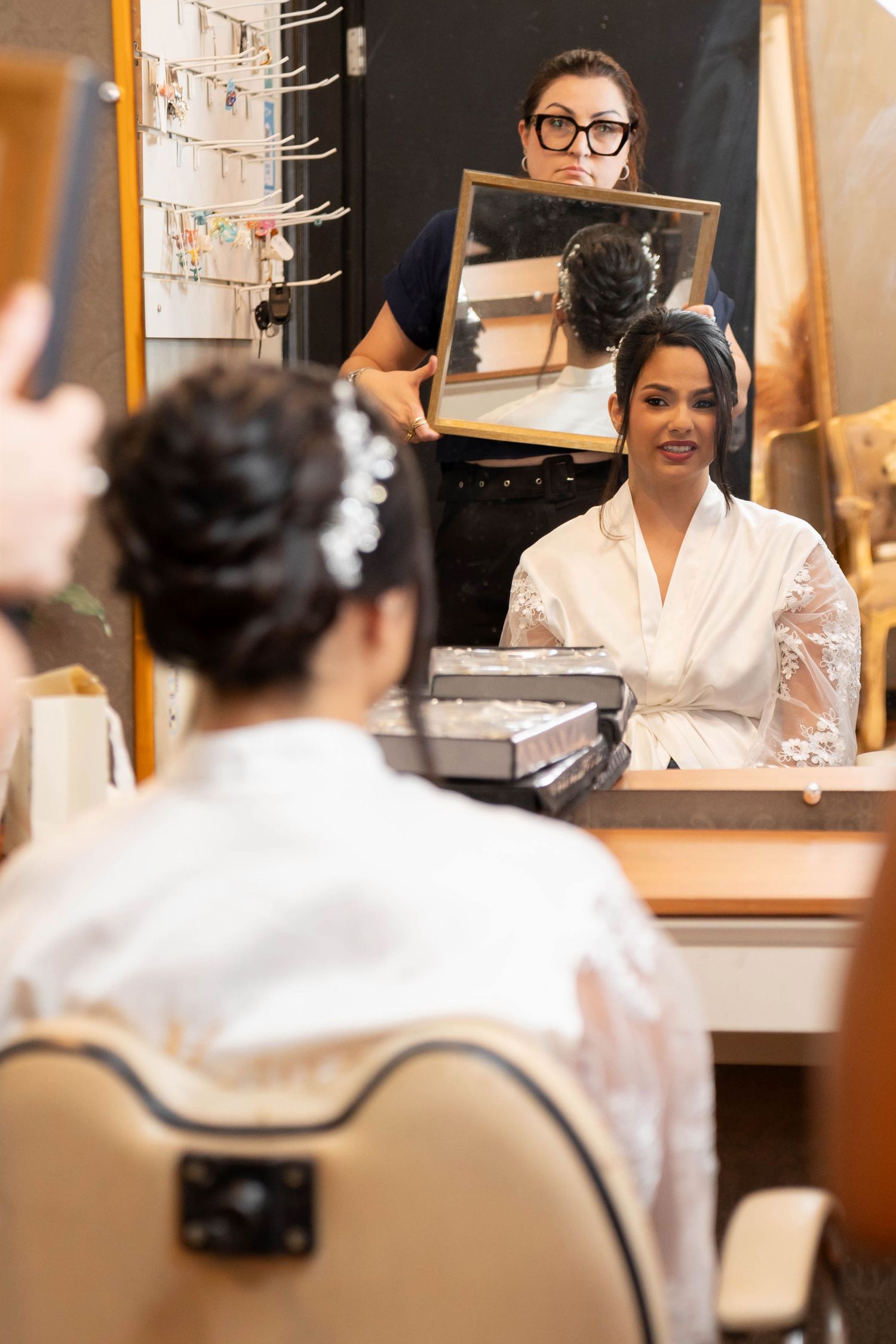 Bride in white robe sits at vanity mirror while hairstylist holds up small mirror to show her elegant updo hairstyle.