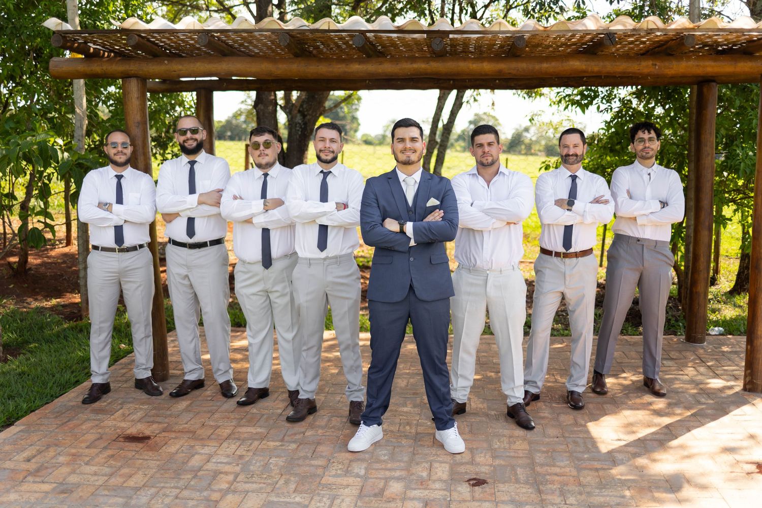 Groom in navy suit poses with groomsmen in white shirts and ties under a wooden pergola at an outdoor wedding.