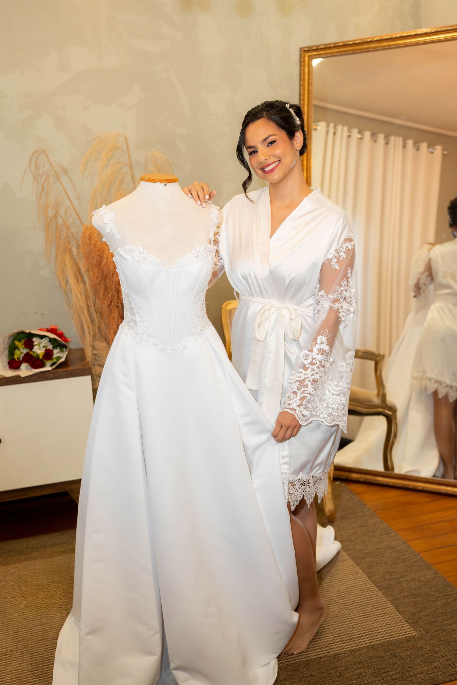 Smiling bride in white lace robe stands beside wedding dress on mannequin in elegant bridal suite.