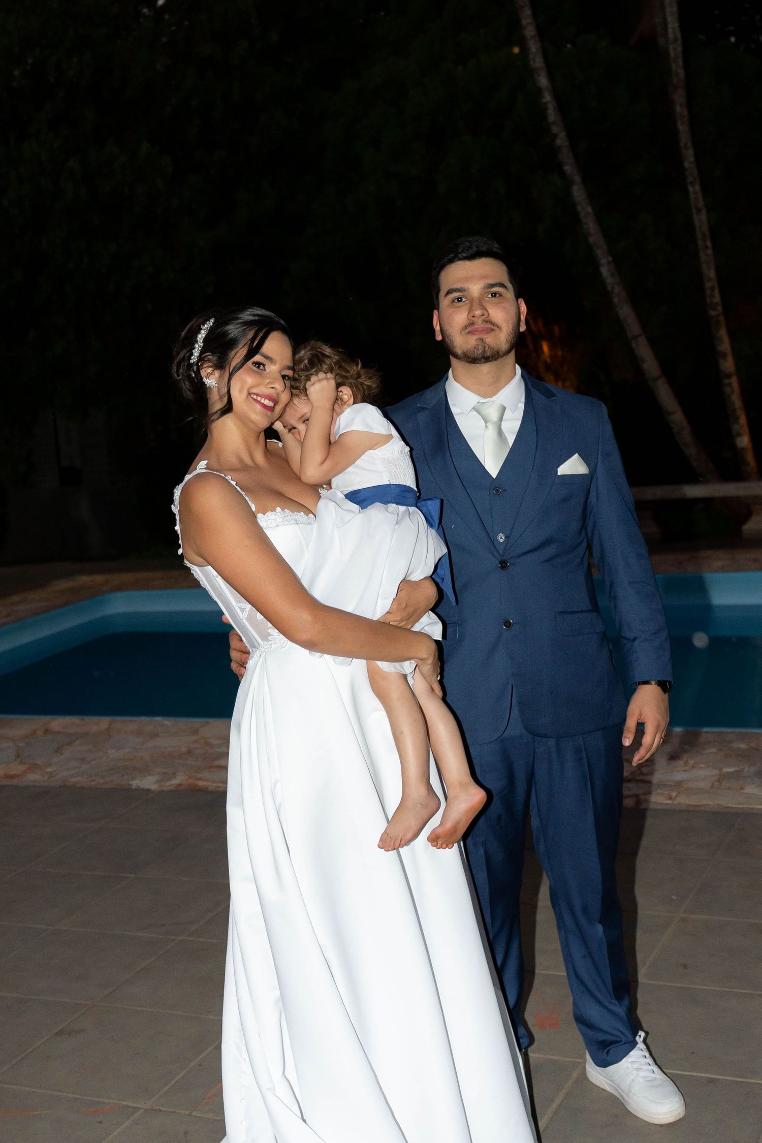 Bride in white gown holds child while groom in blue suit stands beside pool at night wedding celebration.