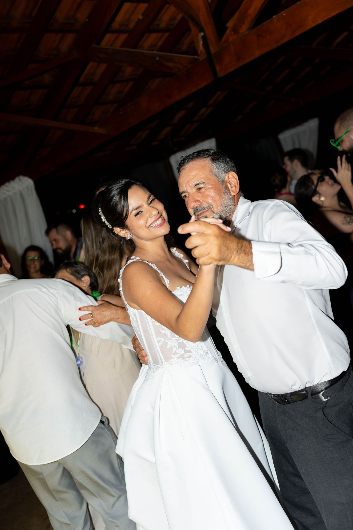 Bride in white lace gown shares a joyful dance with older man at rustic wood-beamed wedding reception.