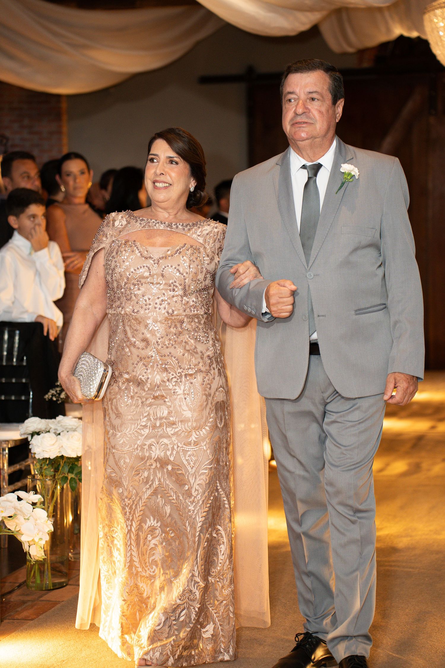 Elegant couple walking down wedding aisle, woman in gold lace gown, man in gray suit with boutonniere.