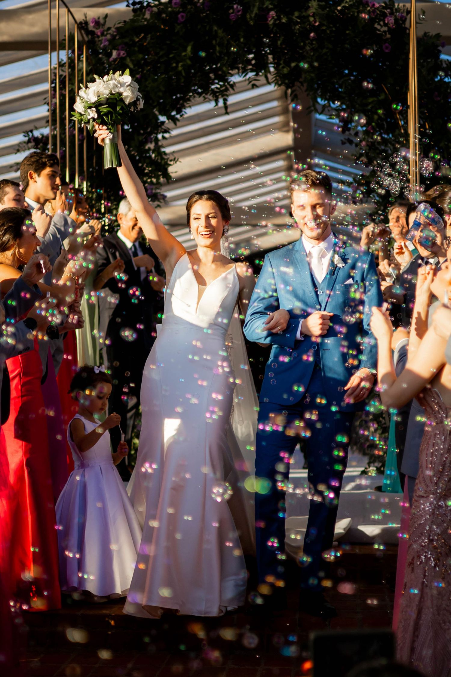 Joyful bride raises bouquet as couple exits ceremony arch surrounded by guests and floating soap bubbles.