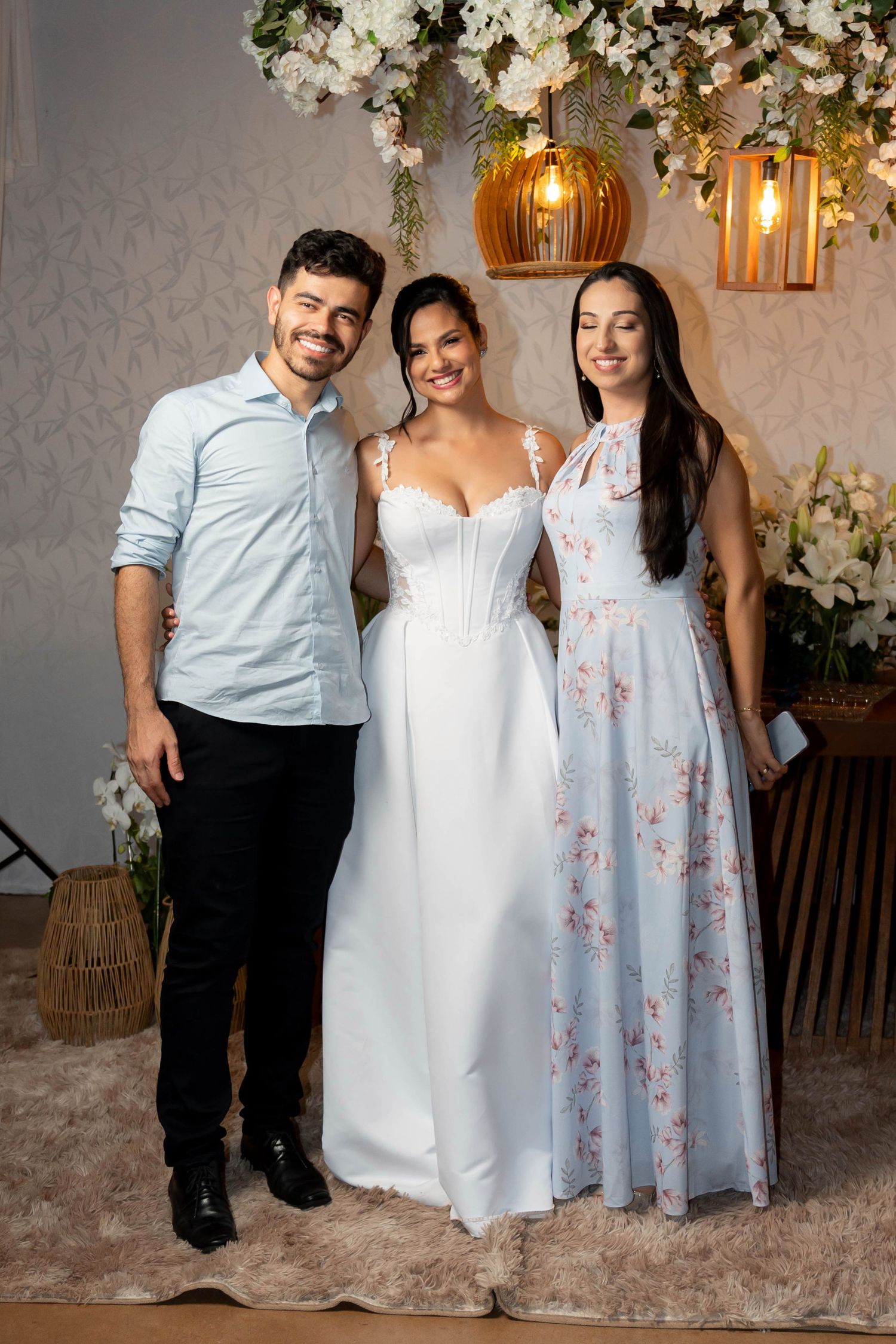 Bride in white gown poses with smiling man and woman in floral dress at decorated wedding venue.