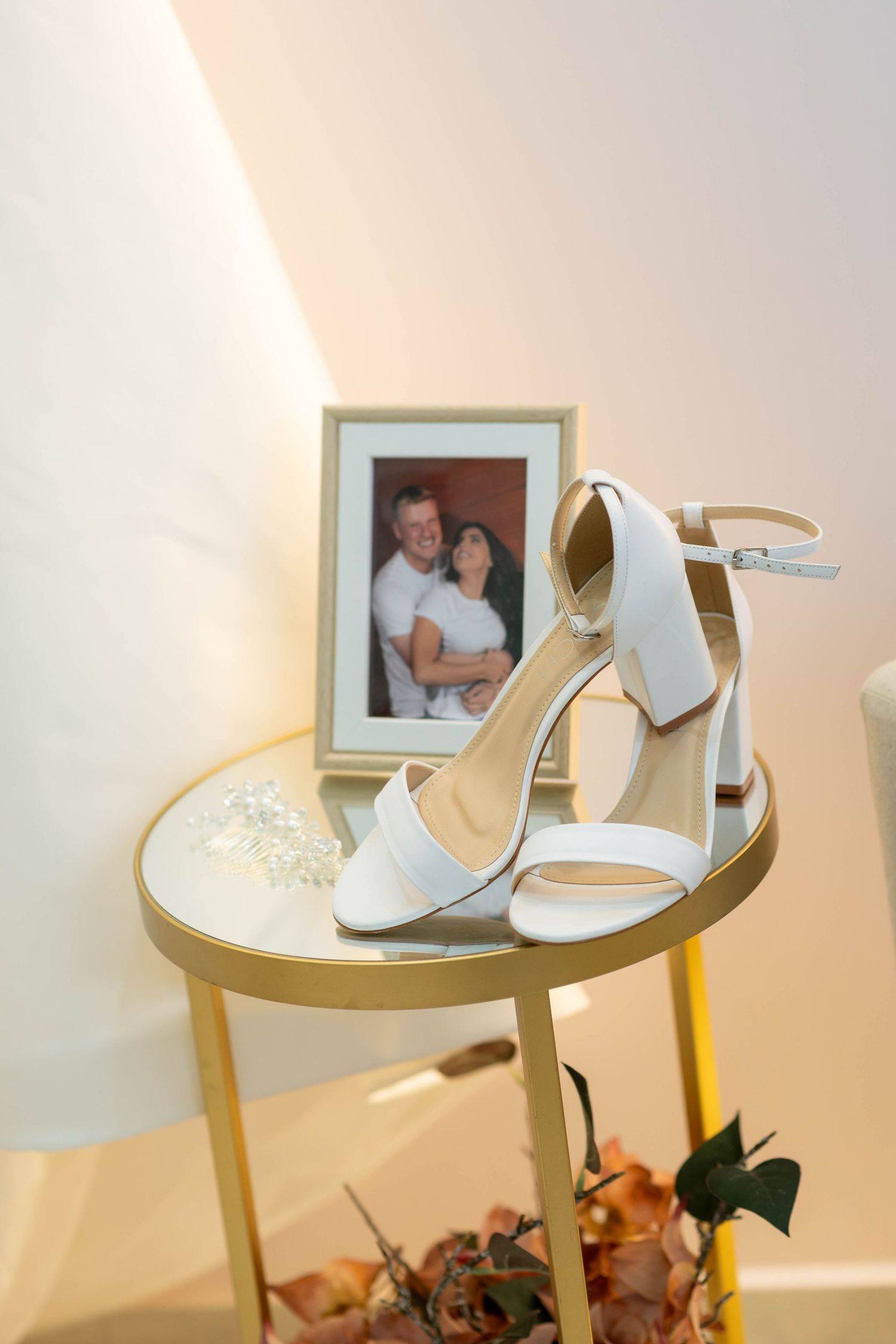 White bridal heels on gold table with couple photo frame and autumn flowers in soft warm light.