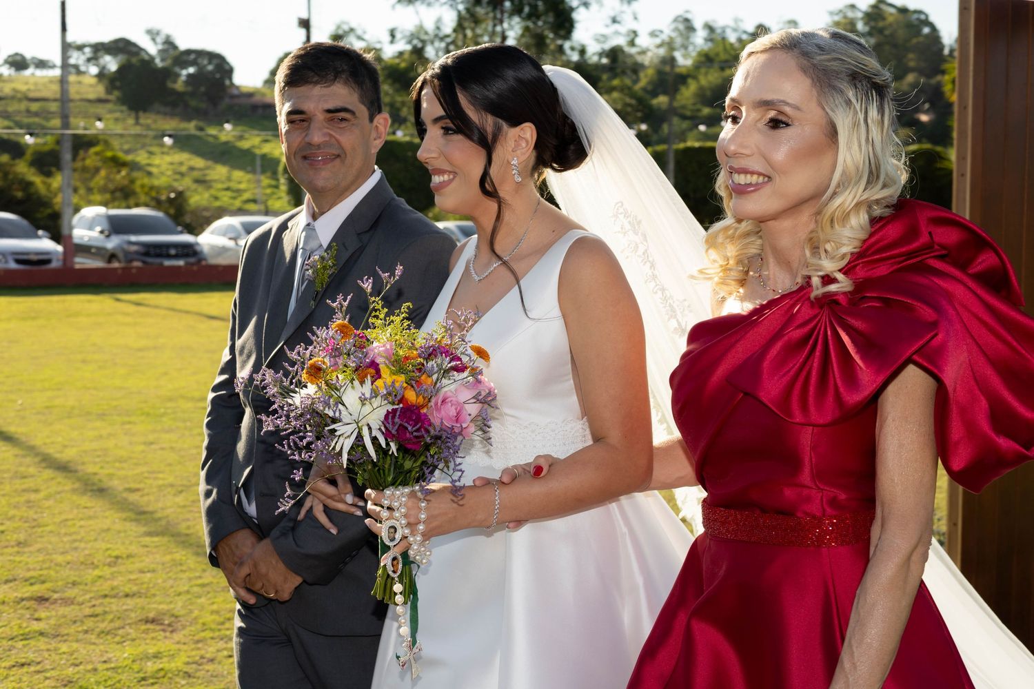 Bride in white gown holding colorful bouquet stands with groom in gray suit and woman in red dress at outdoor wedding.