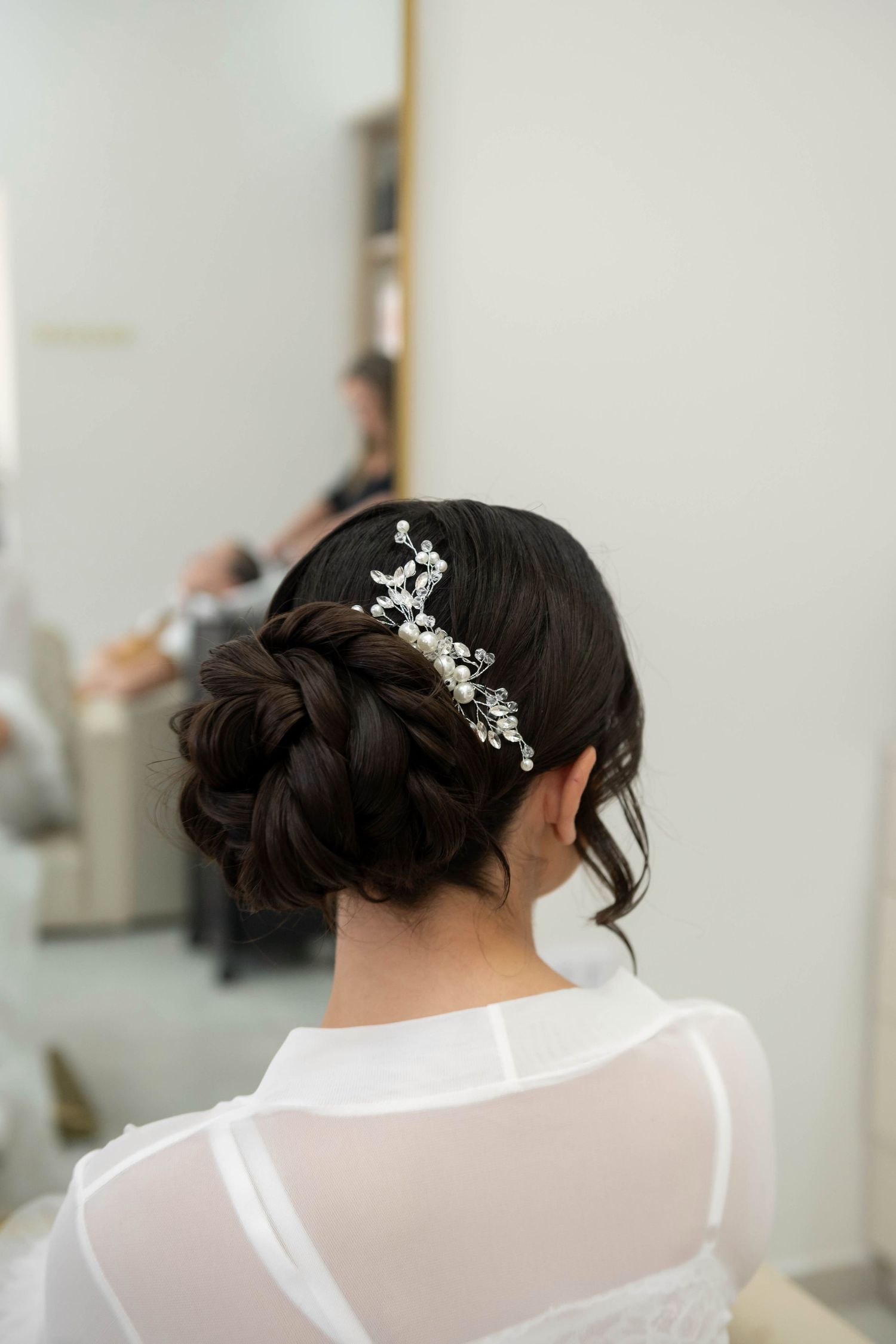 Elegant bridal updo with decorative pearl hairpiece on dark hair, viewed from behind in a salon setting.