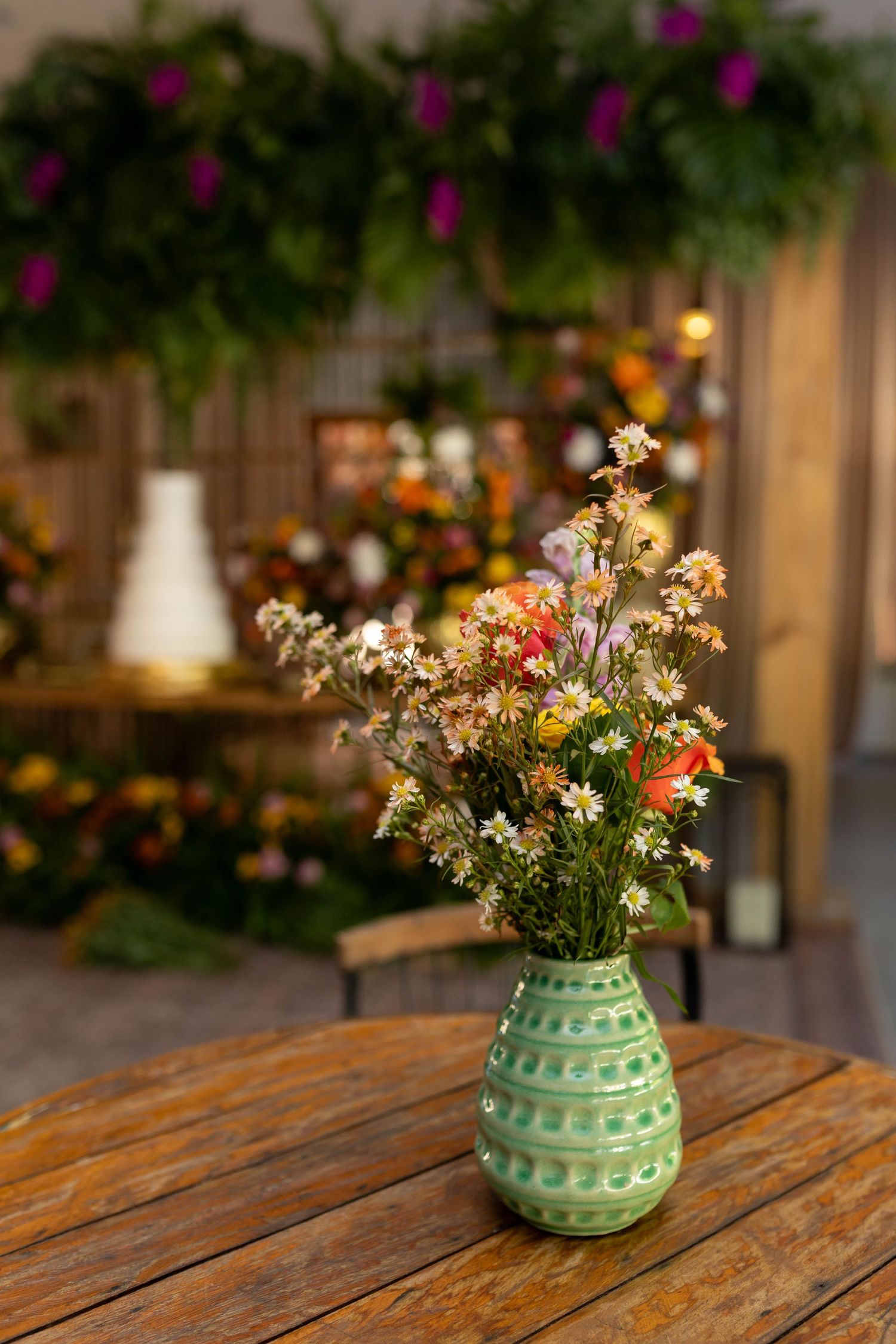 Green ceramic vase with wildflowers and daisies on wooden table in a decorated garden setting.