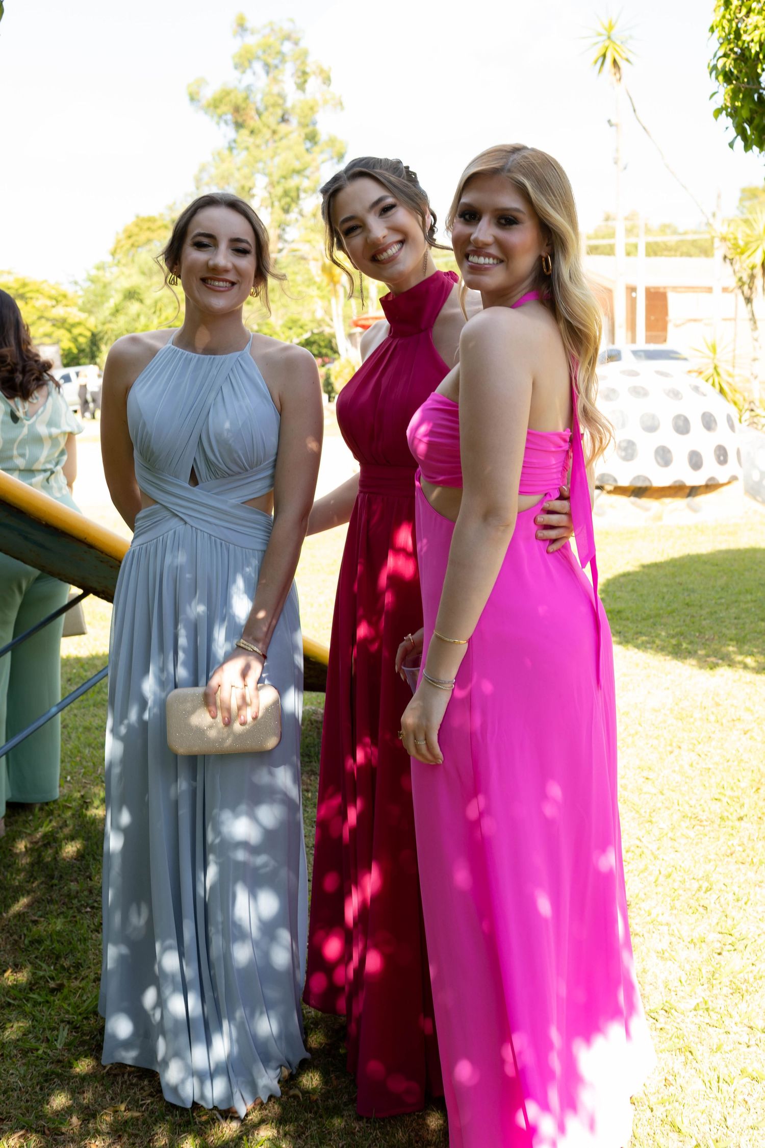 Three young women in elegant formal gowns posing outdoors, wearing light blue, magenta, and pink dresses at a special event.