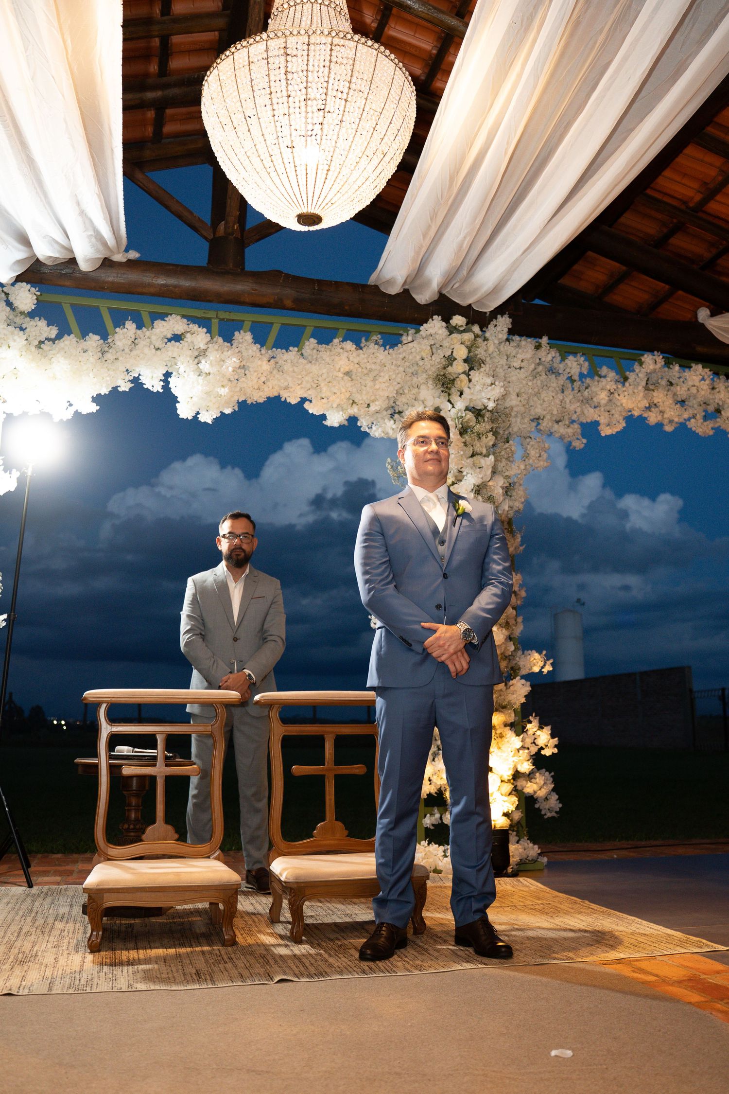Two grooms in suits stand at a floral wedding altar under a chandelier at an elegant outdoor evening ceremony.