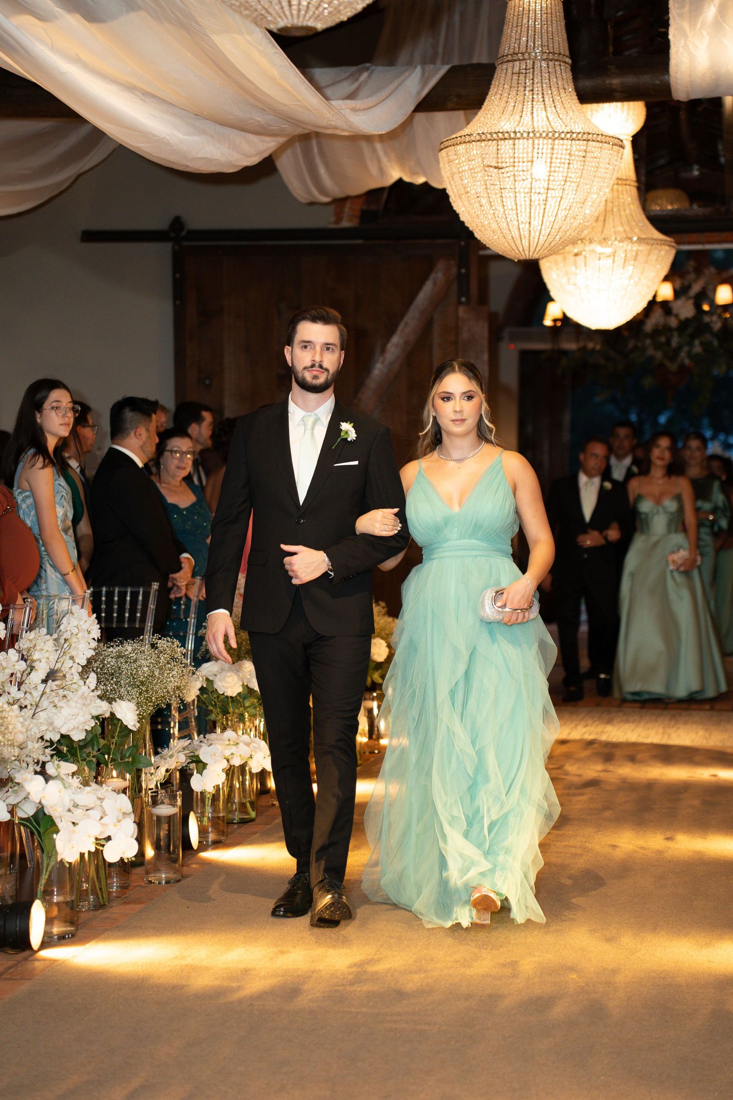 A couple walks down a wedding aisle, man in black tuxedo and woman in mint green gown, under crystal chandeliers.