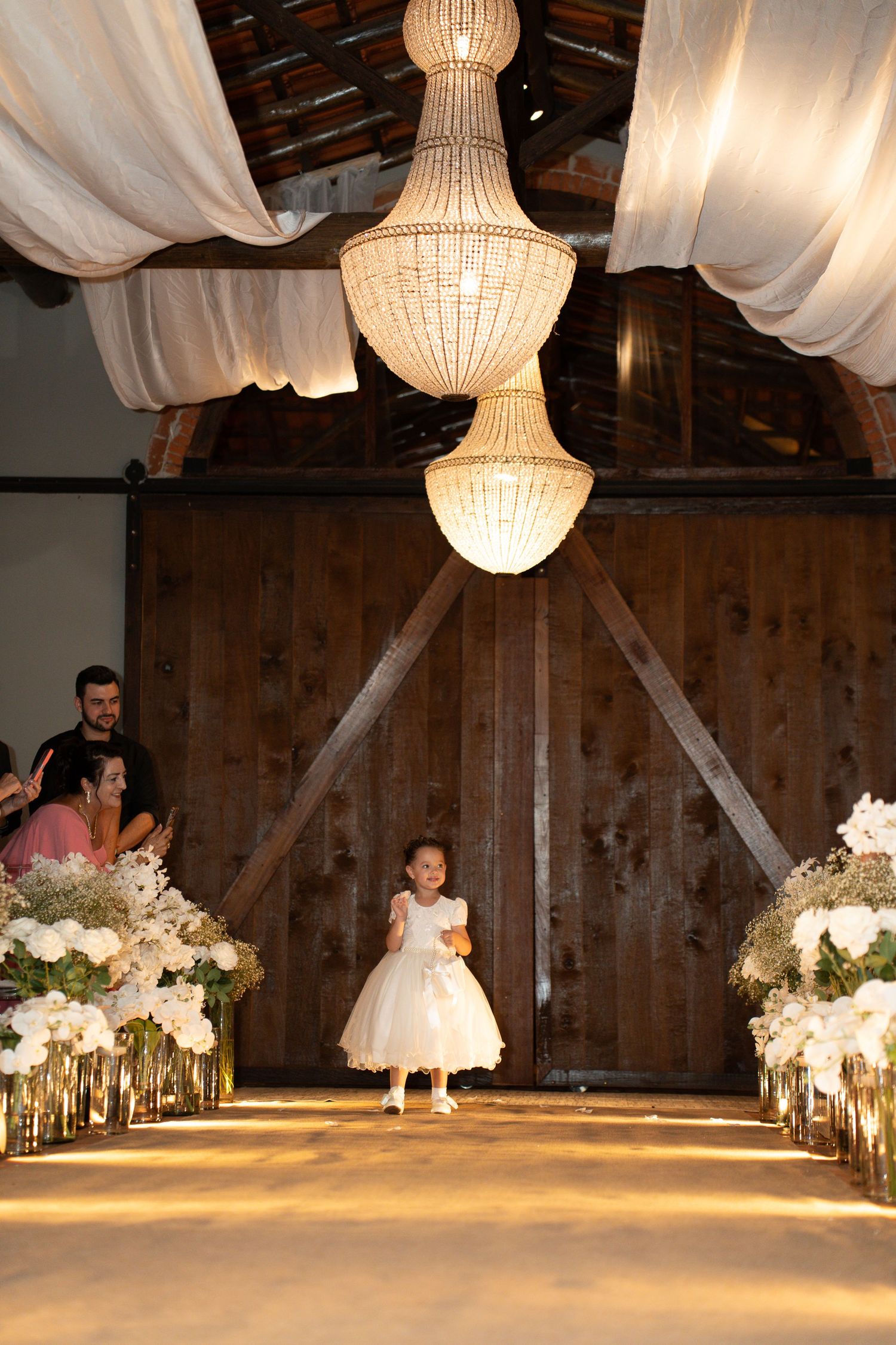 A flower girl in a white dress walks down a decorated wedding aisle in a rustic barn with elegant chandeliers.