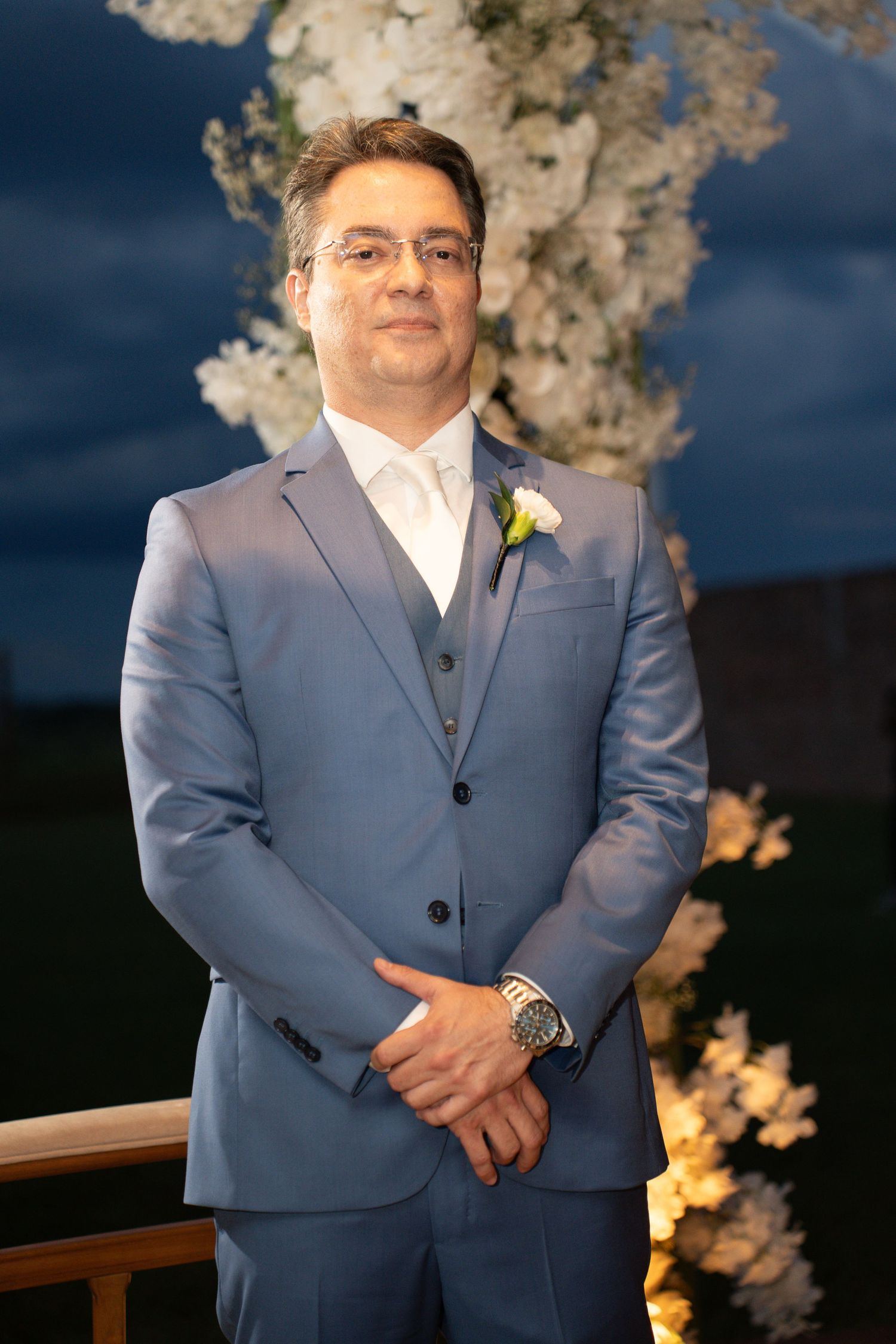 A man in a blue suit with a white boutonniere stands confidently near a floral arch at an evening wedding ceremony.