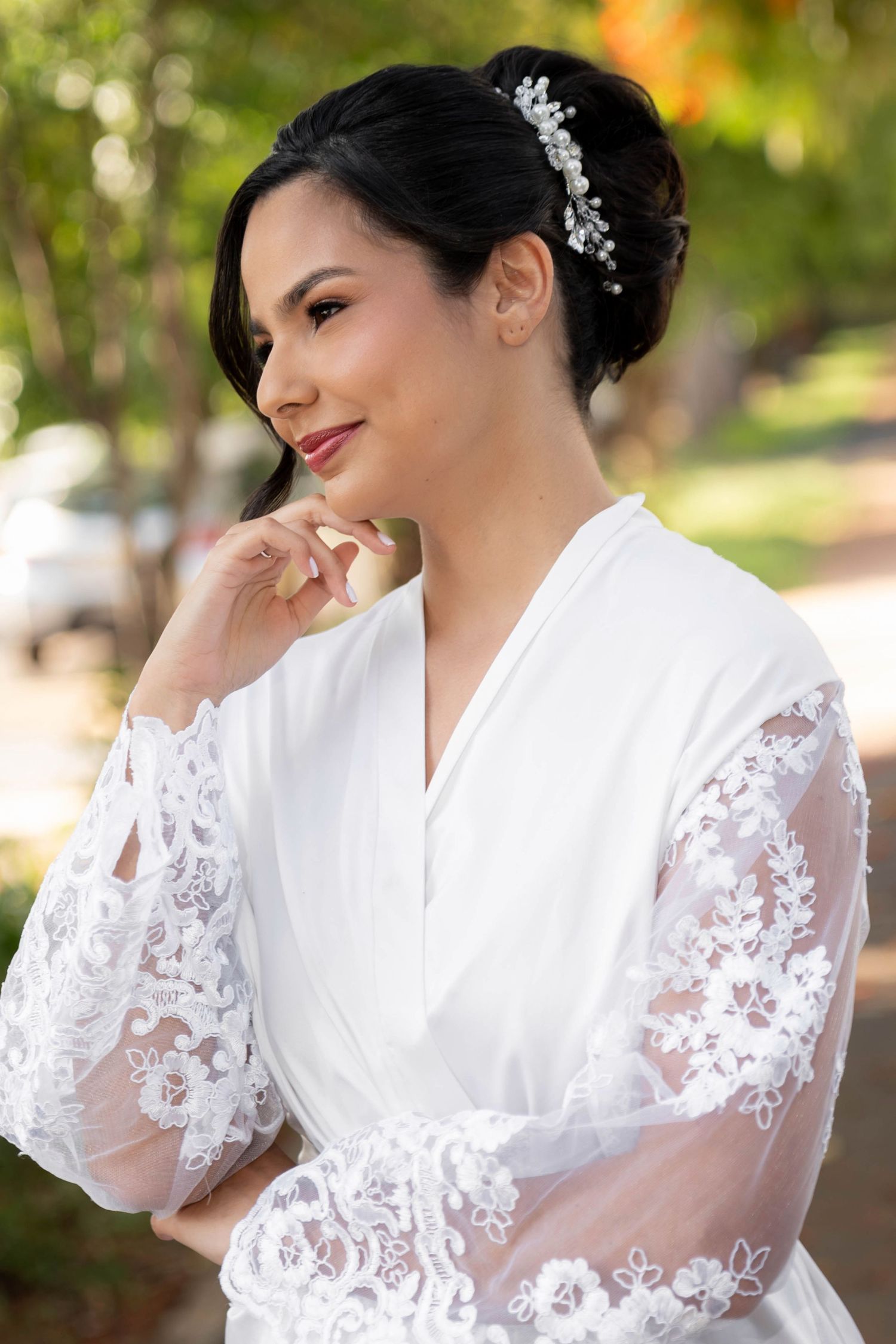 Bride in white lace robe with pearl hair accessory smiling outdoors on her wedding day.