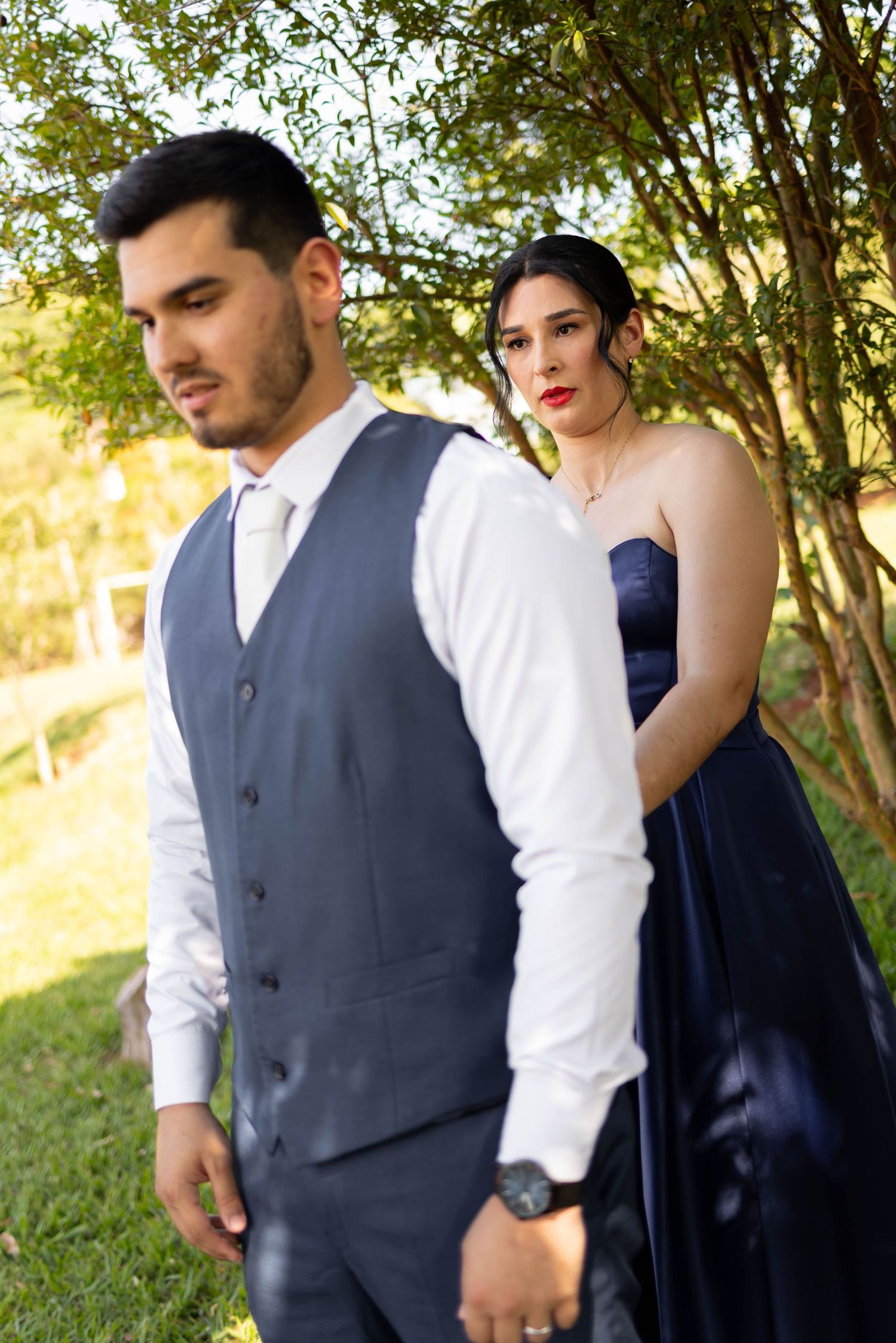 Elegant couple in formal attire outdoors, man in gray vest and woman in navy blue gown posing near green trees.