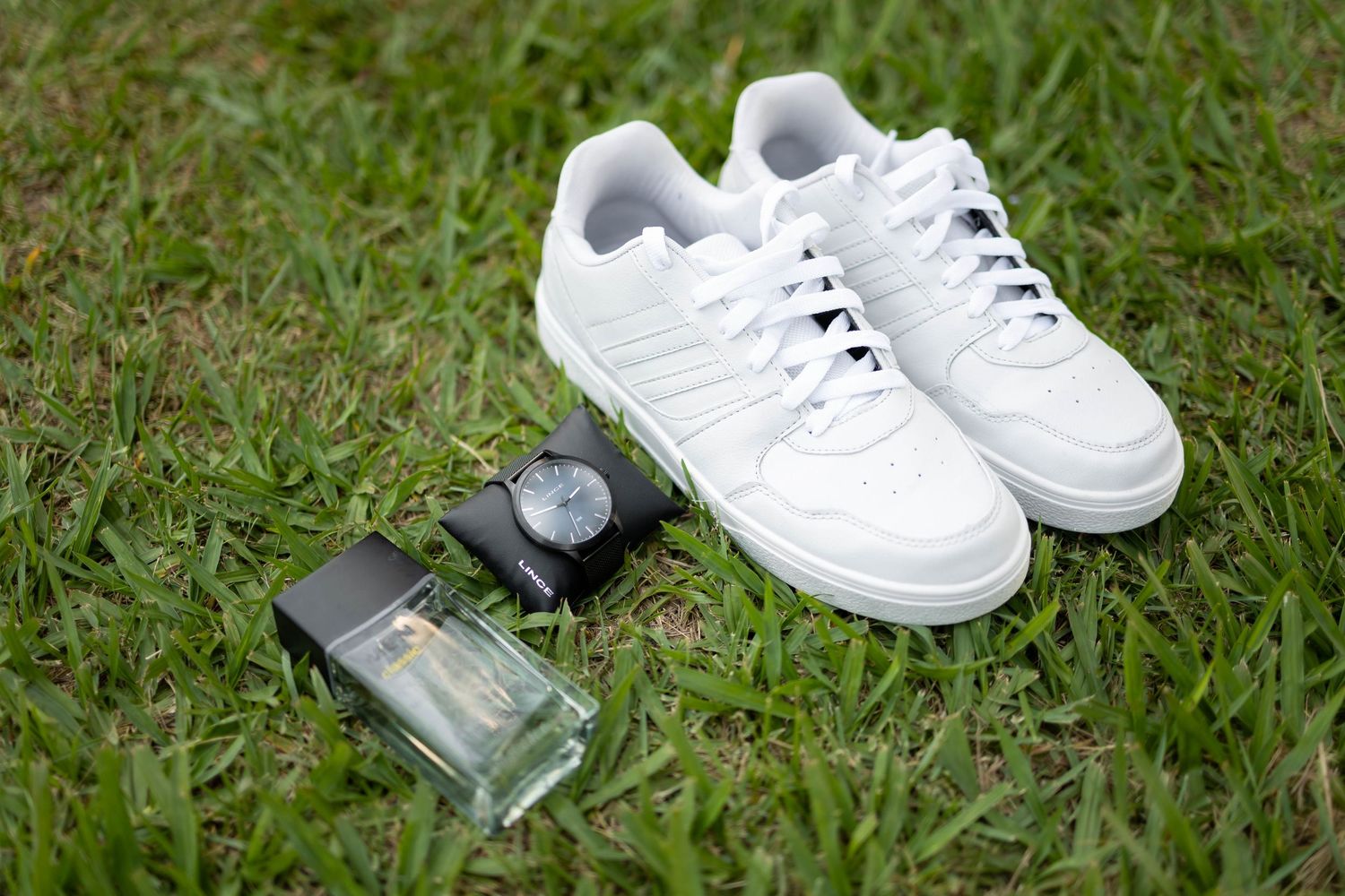 White Adidas sneakers, a black wristwatch, and cologne bottle arranged on green grass outdoors.