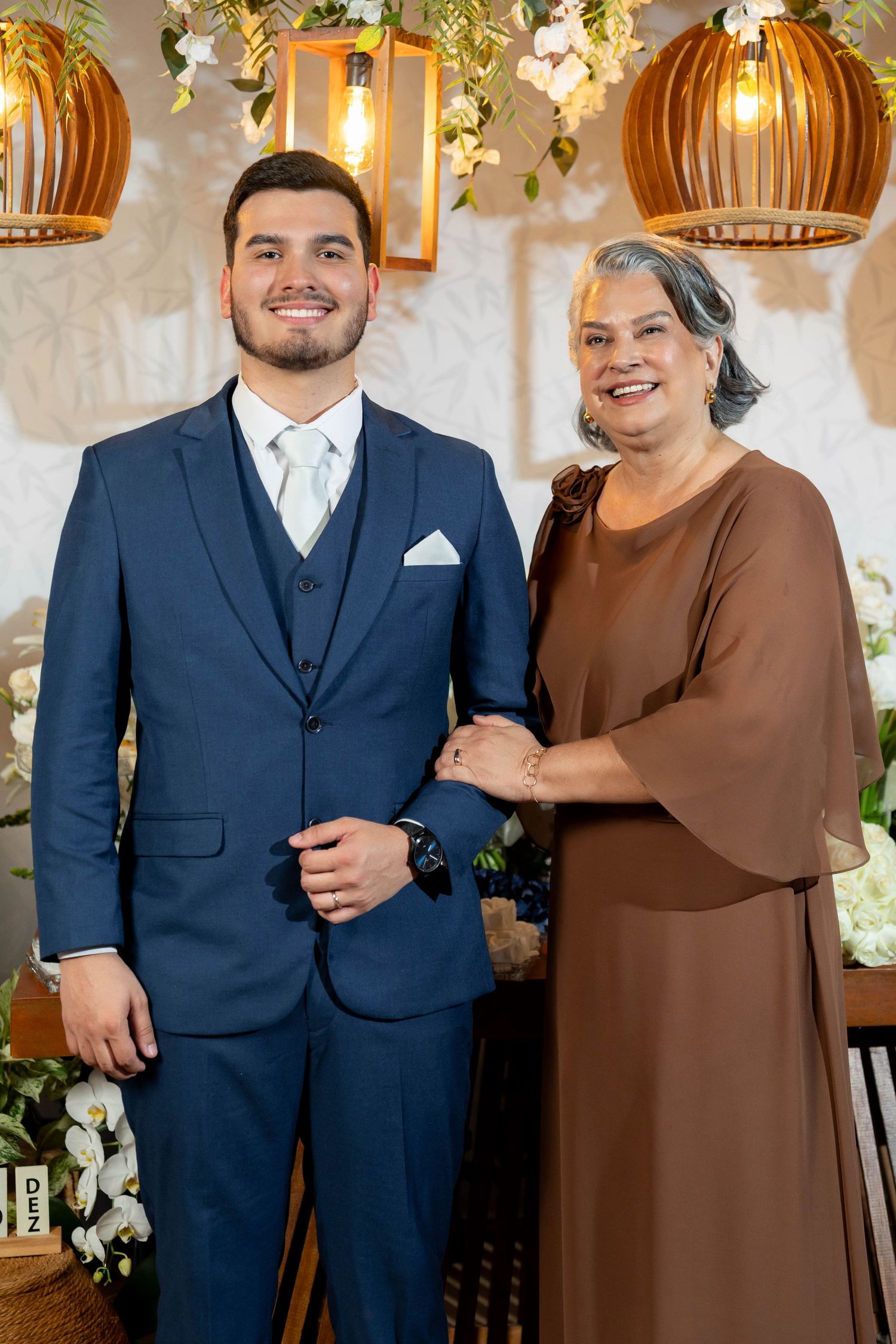 Young man in navy suit poses with older woman in brown dress at elegant wedding venue with warm lighting.