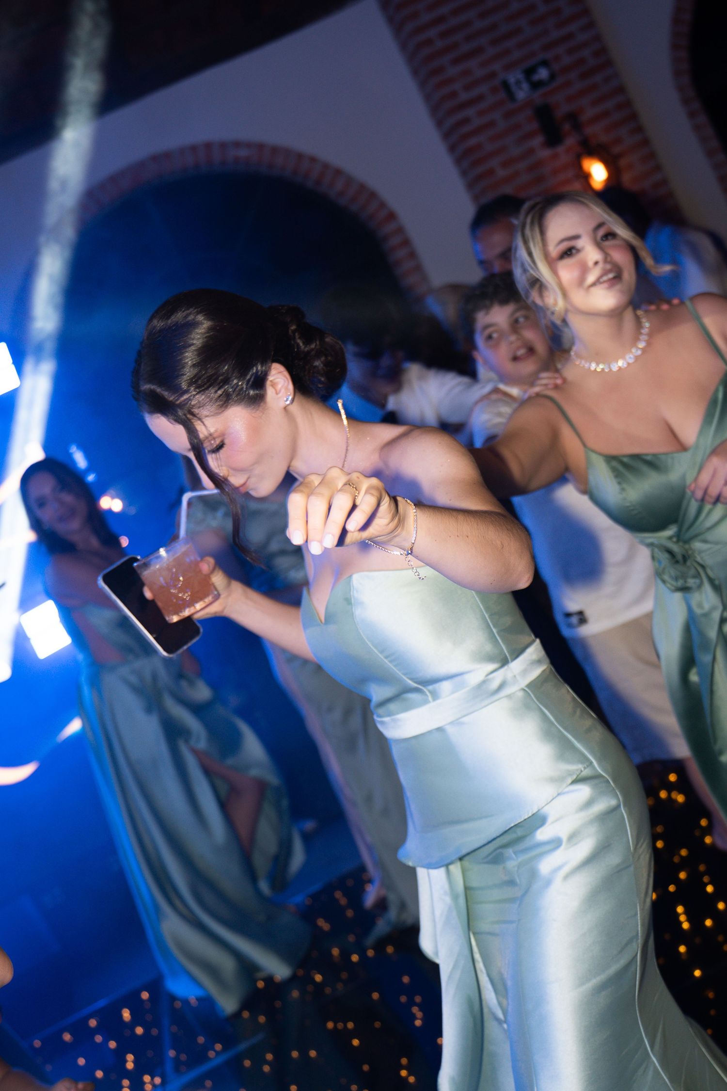 Bridesmaids in sage green satin dresses dancing energetically at a wedding reception under blue lights.