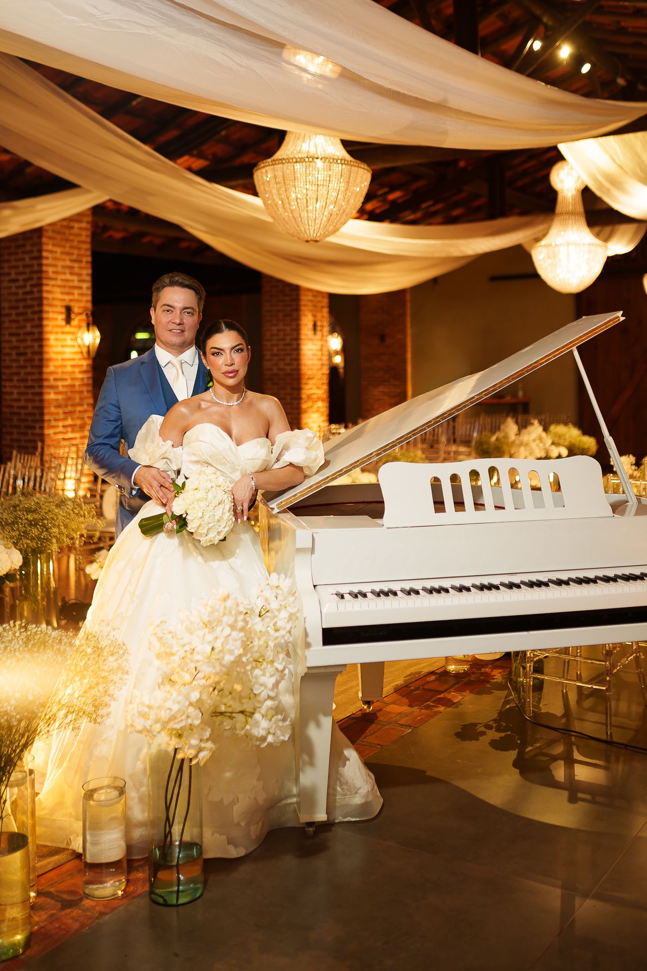 Elegant bride and groom posing beside a white grand piano in a warmly lit wedding venue with draped ceilings.