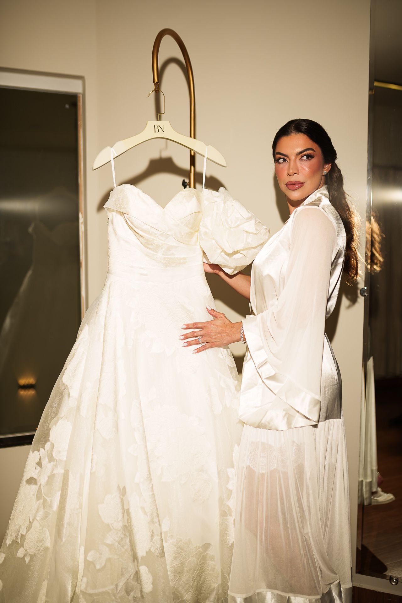 Woman in white satin robe admiring an elegant strapless lace wedding dress on a gold hanger in a bridal boutique.