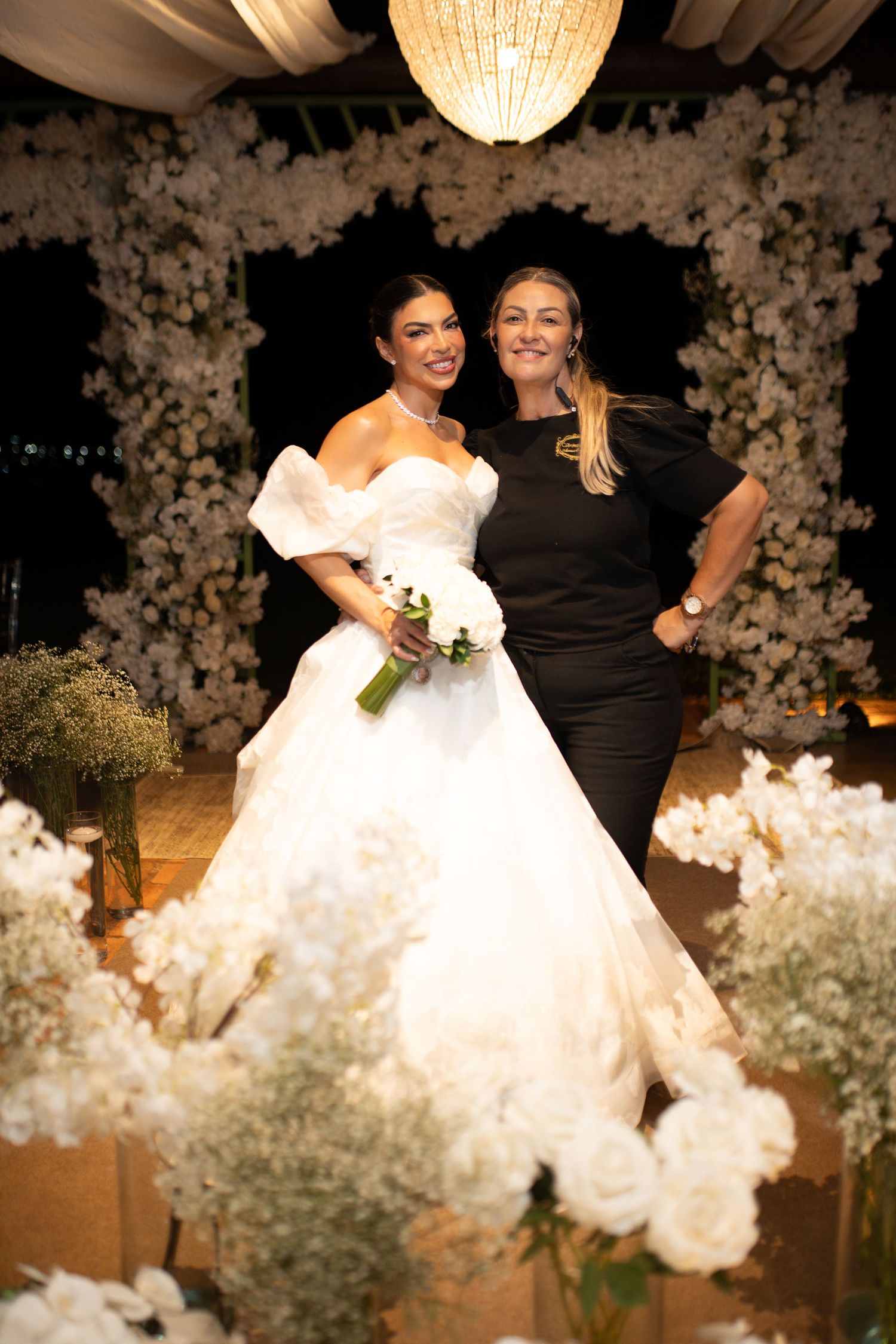 Bride in white off-shoulder ball gown holding bouquet poses with woman in black dress among white floral arrangements.