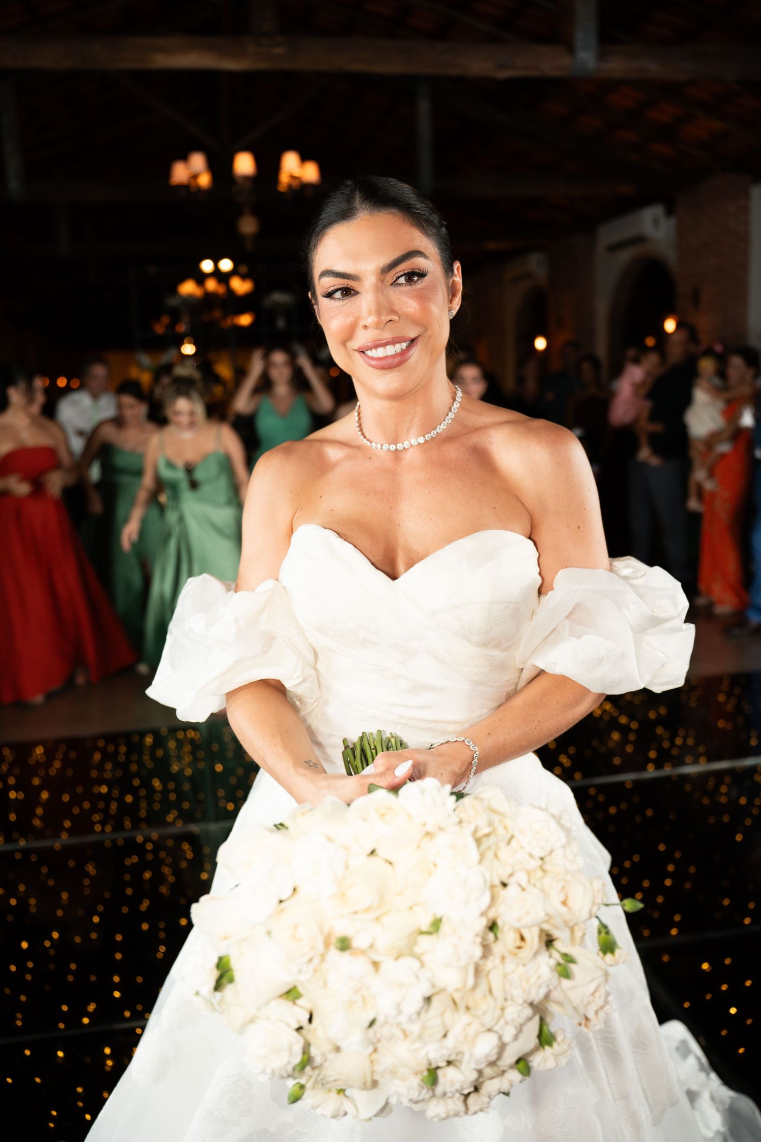 Smiling bride in white strapless gown holding large white floral bouquet at elegant wedding reception.