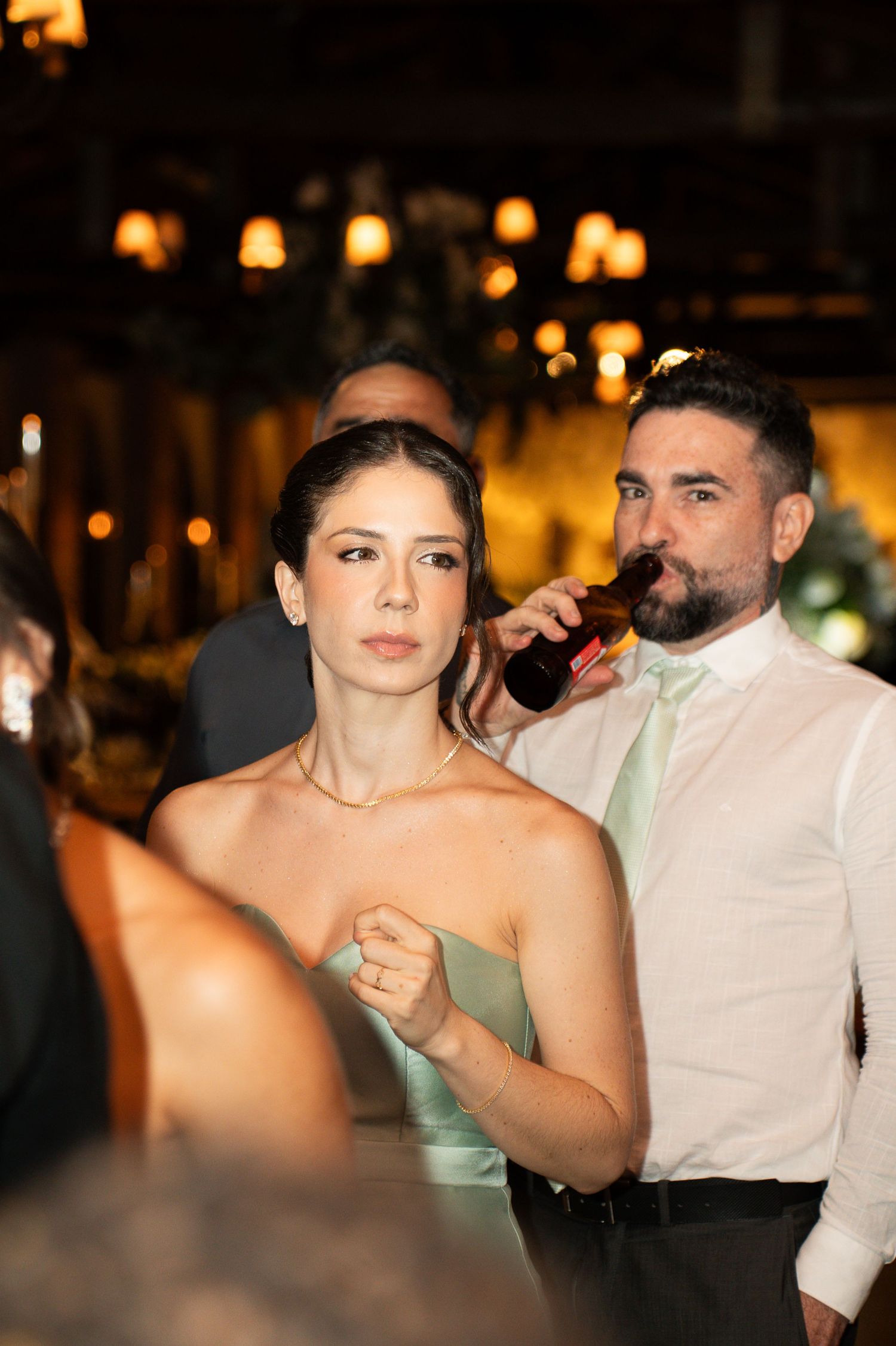 A woman in a sage green dress and a bearded man in a white shirt drinking beer at an elegant evening wedding reception.