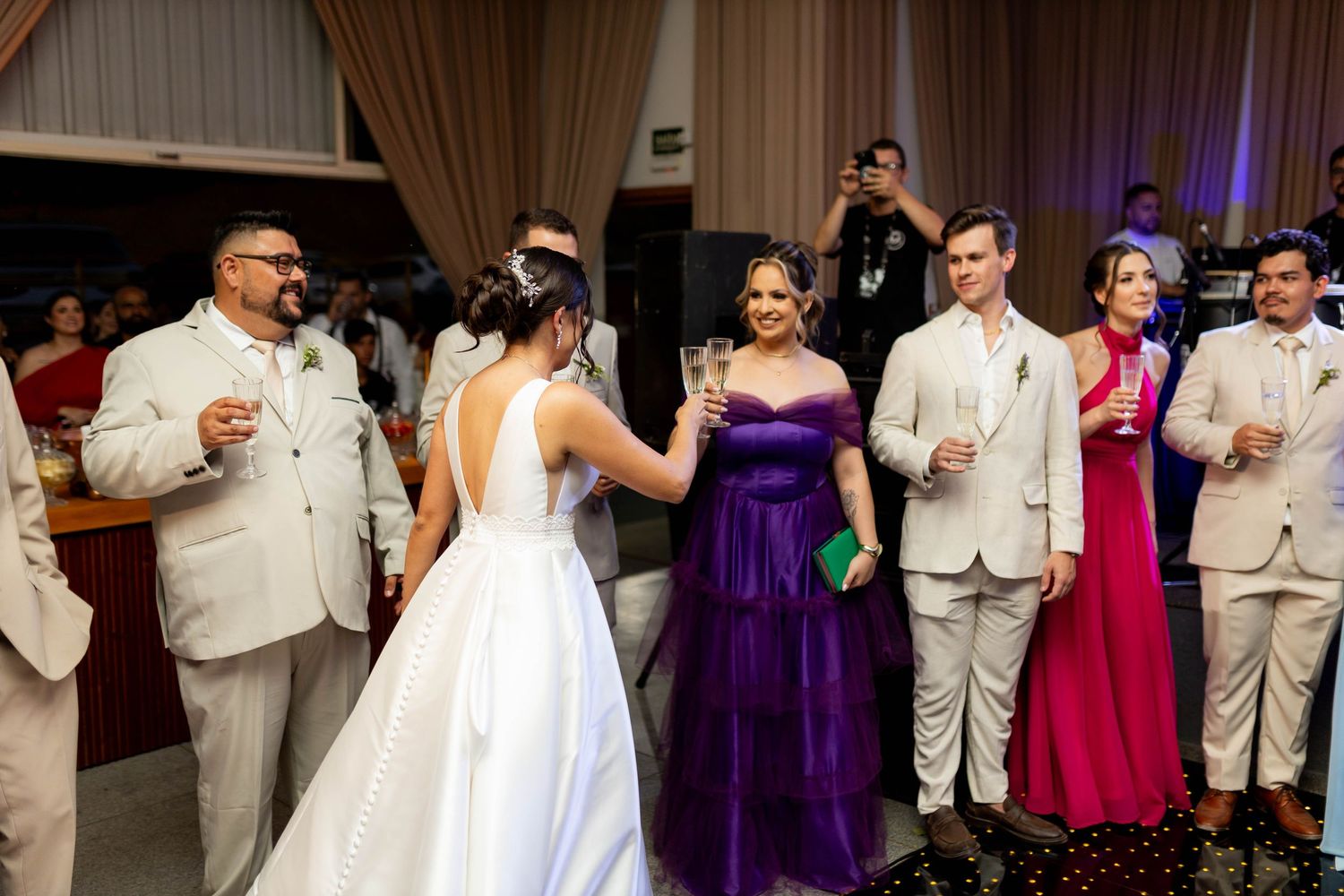 Wedding reception guests in formal attire, including bride in white gown and woman in purple dress, socializing indoors.