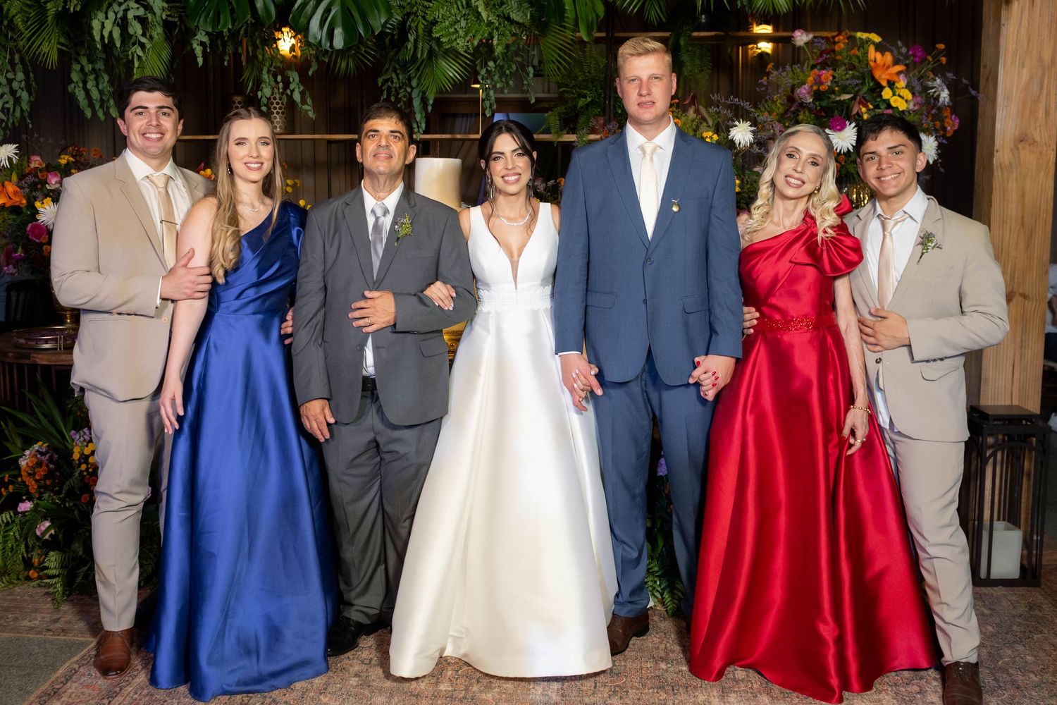 Wedding party of seven posing together, bride in white gown, guests in blue, red, and tan formal attire.