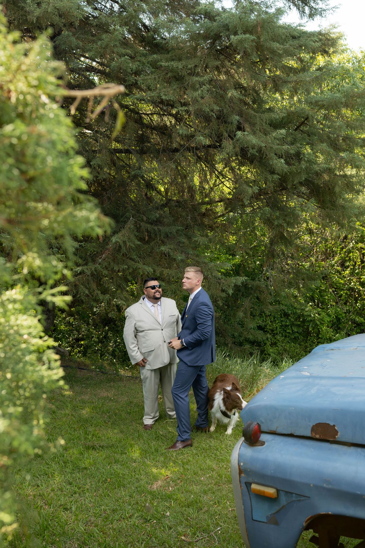 Two men in suits stand on grass near vintage truck and dog, surrounded by lush green trees outdoors.
