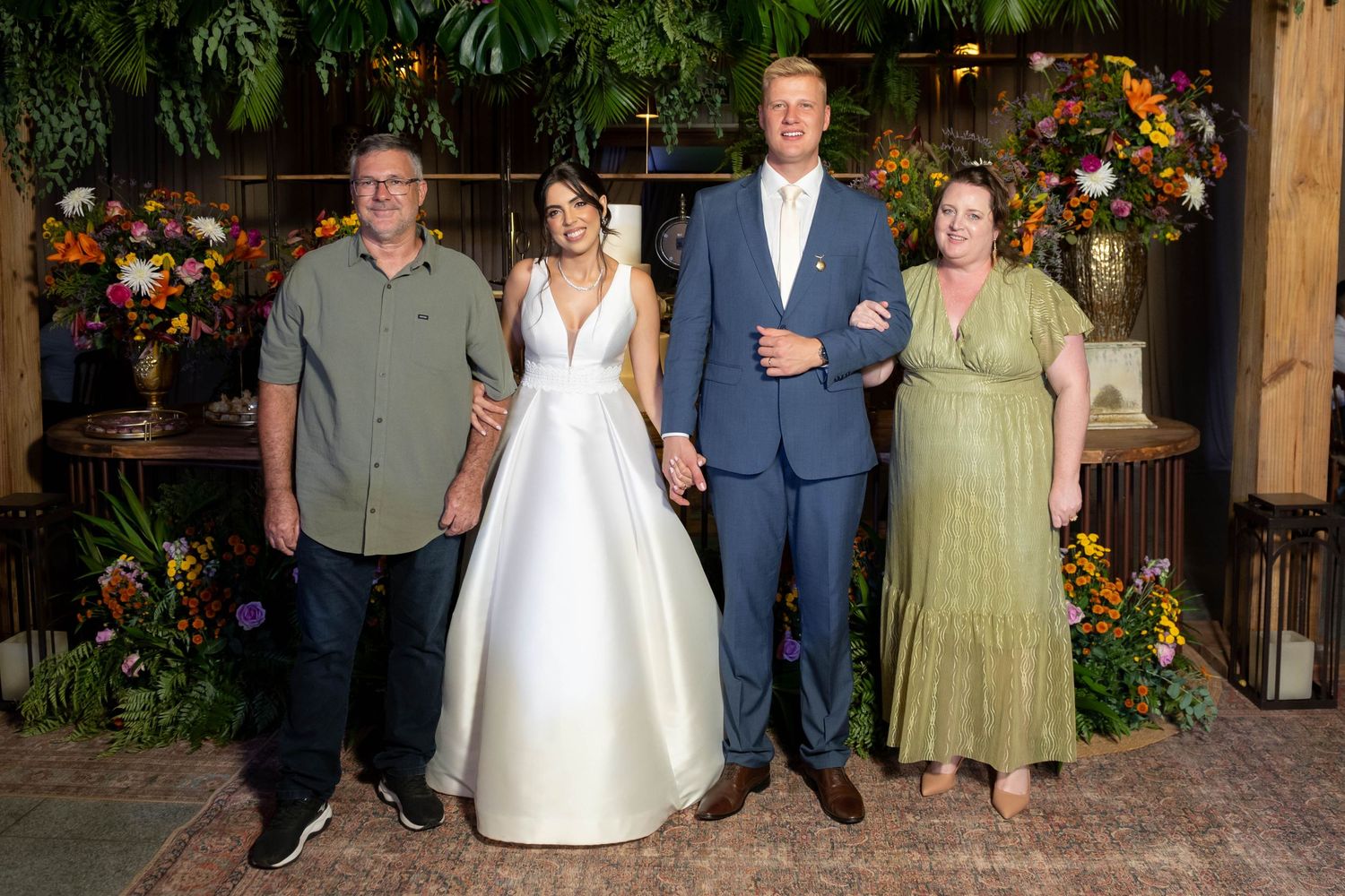 Bride in white gown and groom in blue suit pose with two guests before floral backdrop at elegant wedding reception.