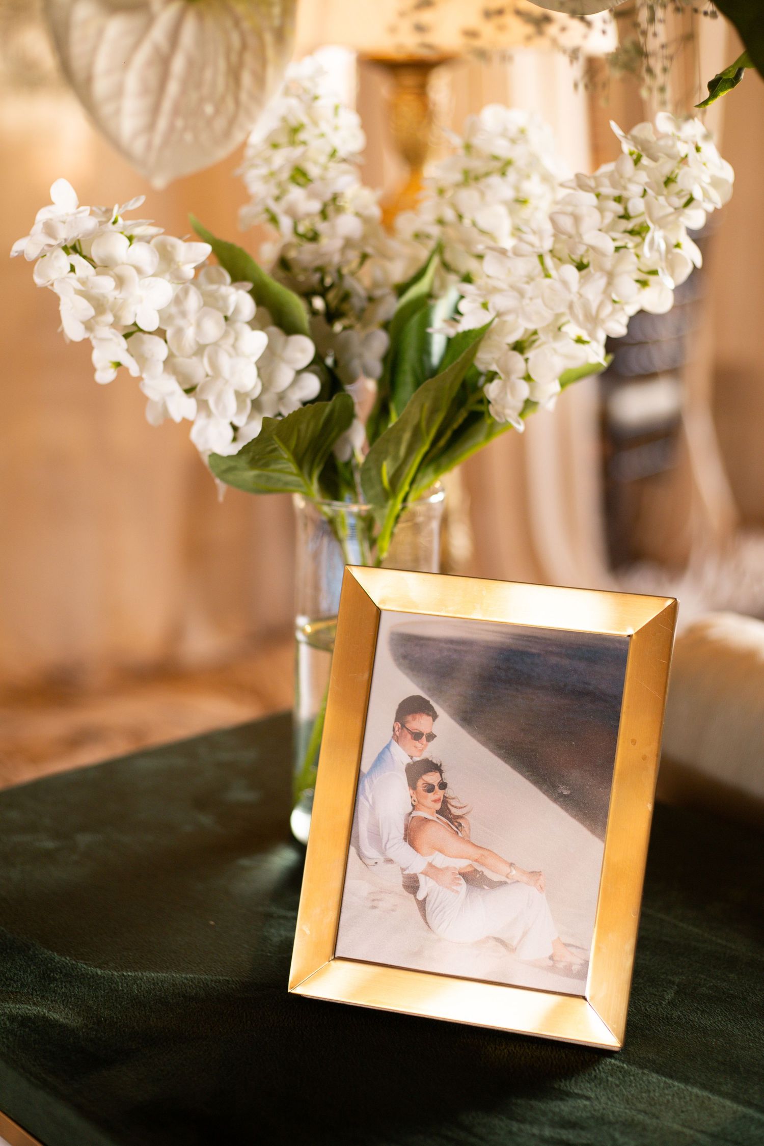 Gold-framed wedding photo beside white hyacinth flowers in a glass vase on a dark green velvet surface.